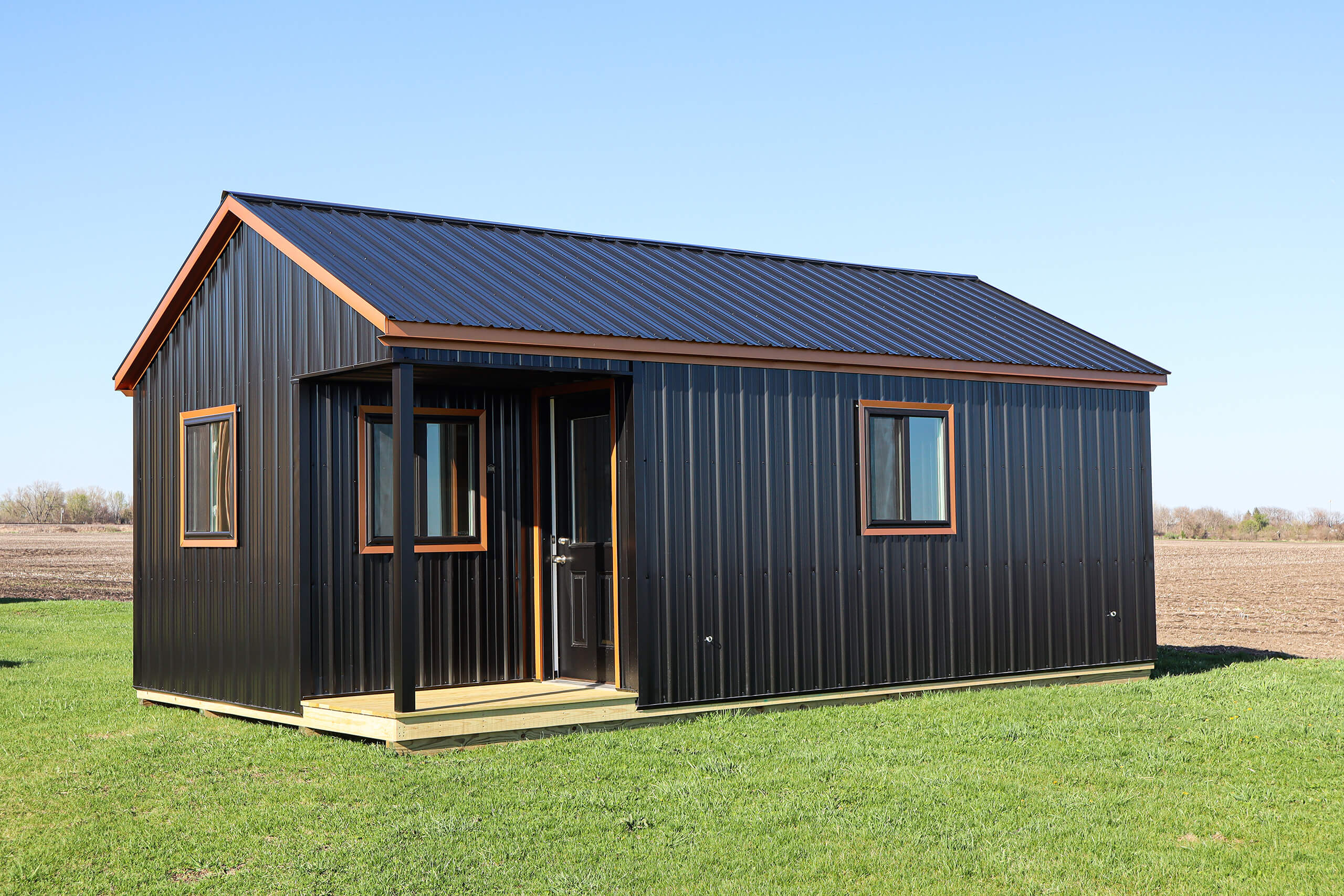 A small, black metal-sided house with a pitched roof and a front porch, situated on a grassy field under a clear blue sky.