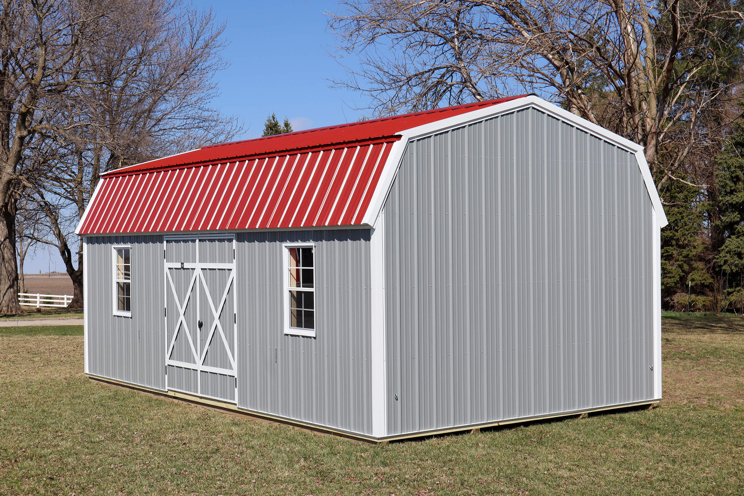 Gray metal storage shed with red roof and double doors, situated on a grassy lawn with bare trees in the background.