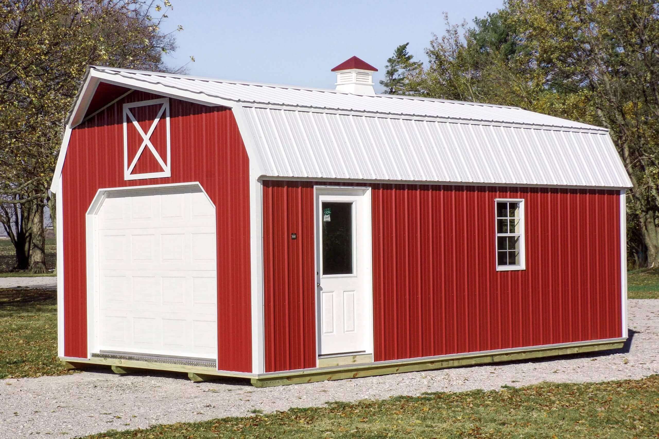 Red barn-style shed with white trim and a small cupola, featuring a large garage door and a white entry door, set on a gravel surface with grass and trees in the background.