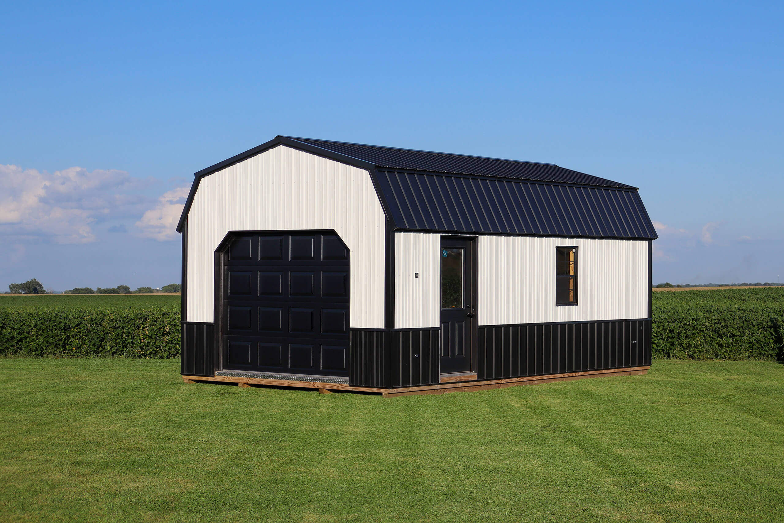 A small black and white metal storage shed with a garage door and single window sits on a grassy lawn with farmland in the background.