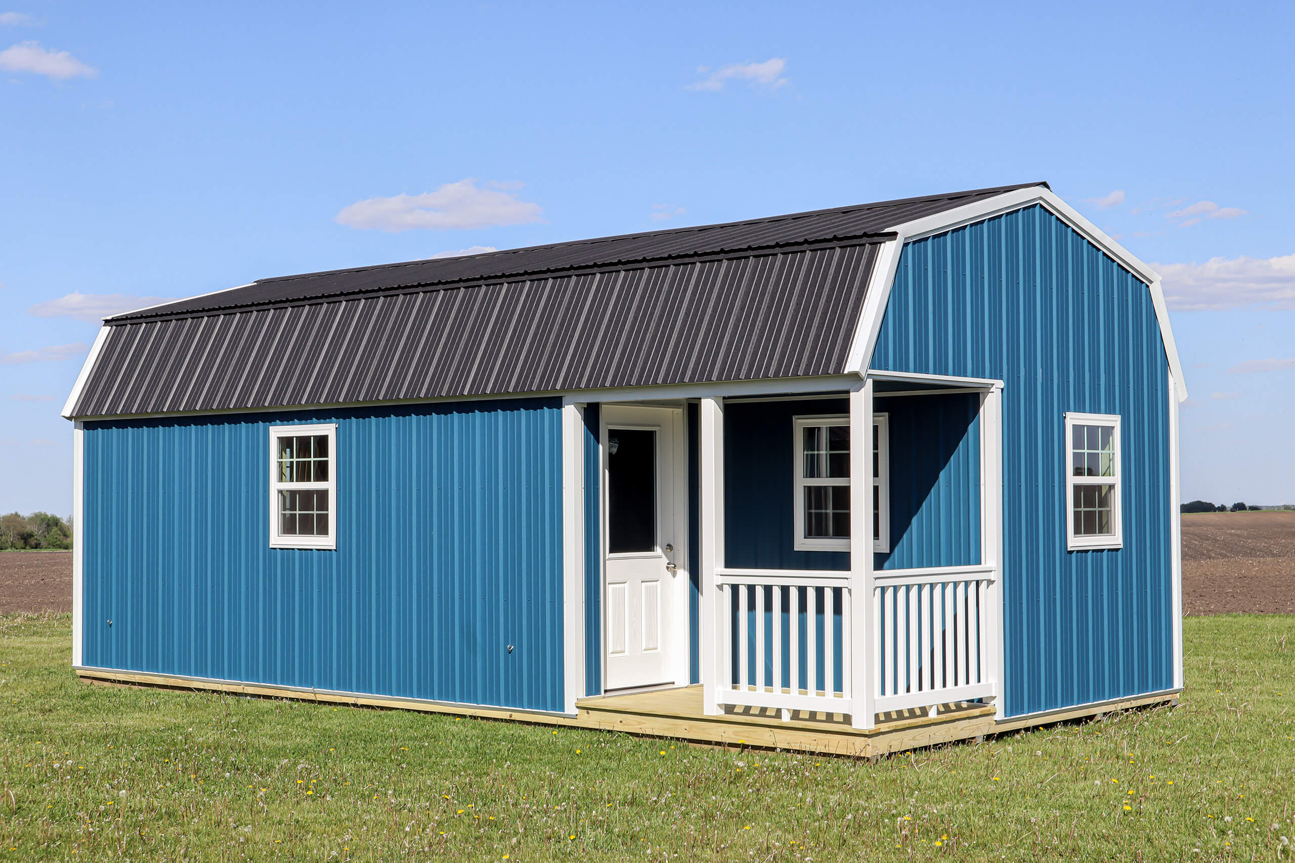 Small blue shed with white trim and a black metal roof, featuring a porch, sits on green grass with an open field in the background under a blue sky.
