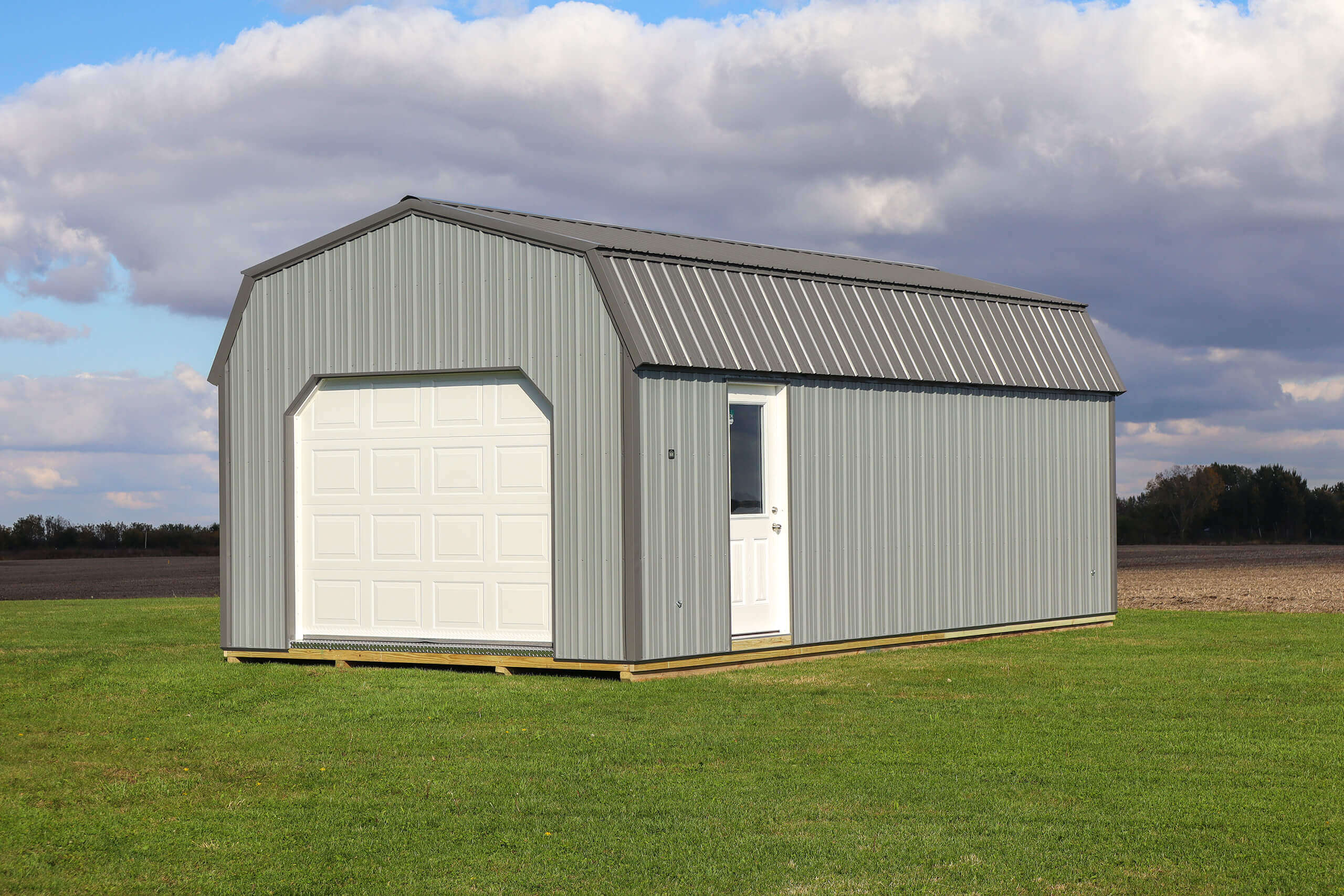 Gray metal storage shed with a white garage door and side entry door, situated on a grassy field under a partly cloudy sky.