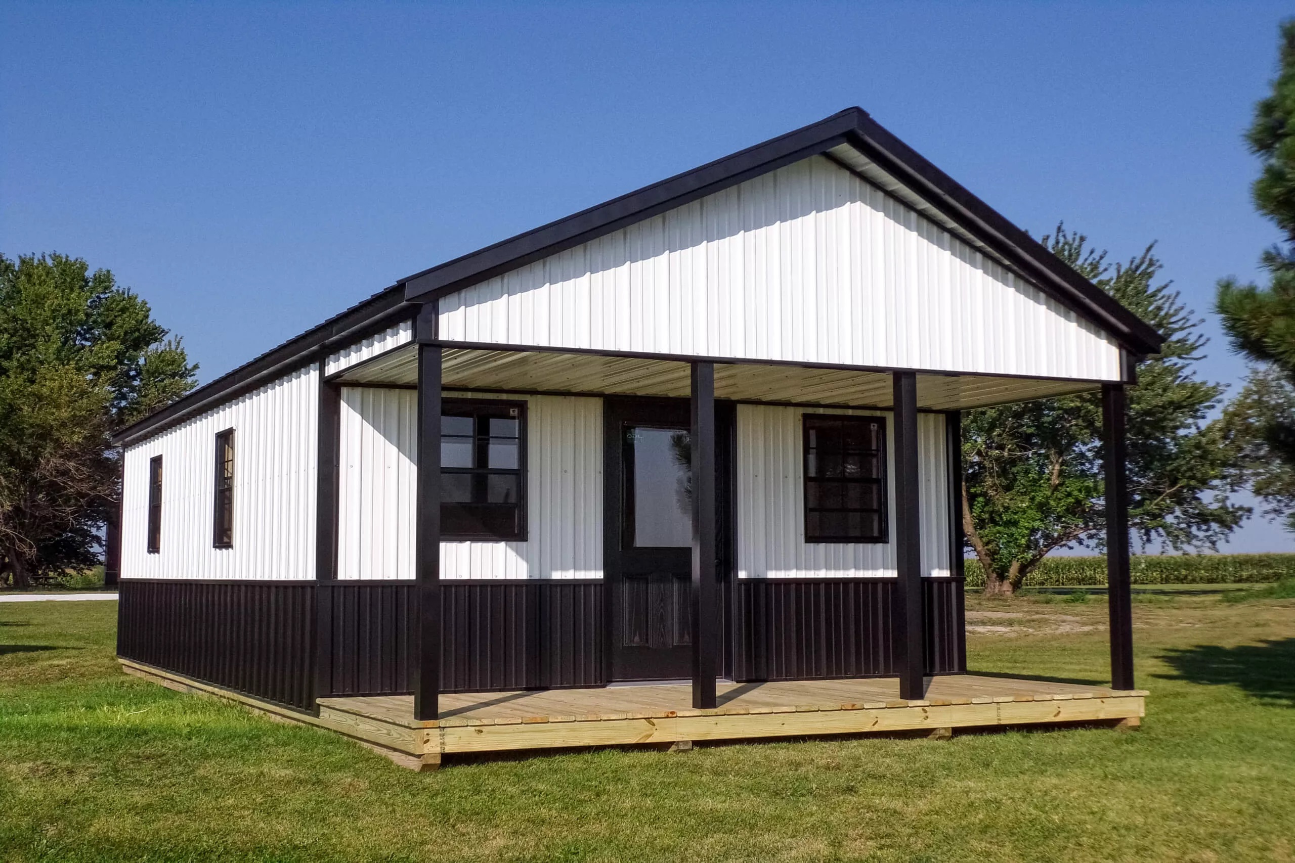 A small, white and black metal building with a front porch stands on a grassy lawn, with trees and clear blue sky in the background.