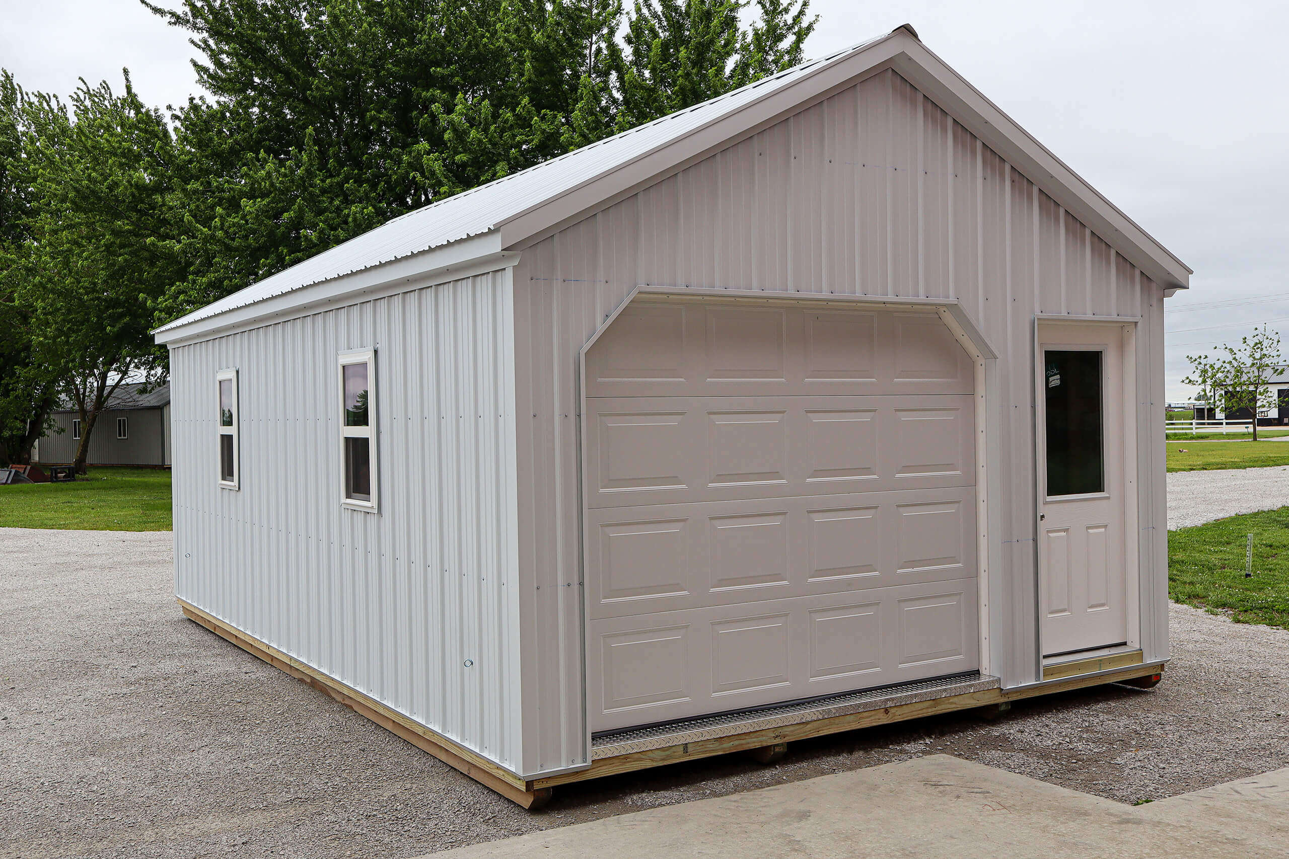 Small white metal shed with a garage door, side window, single entry door, and pitched roof, placed on gravel with grass and trees in the background.