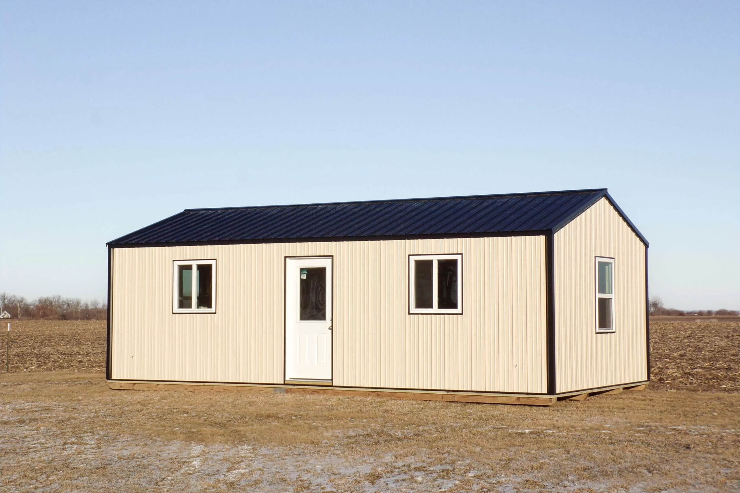 A beige metal portable building with a black roof, two windows, and a white door, situated on dry grass in a flat, open landscape.