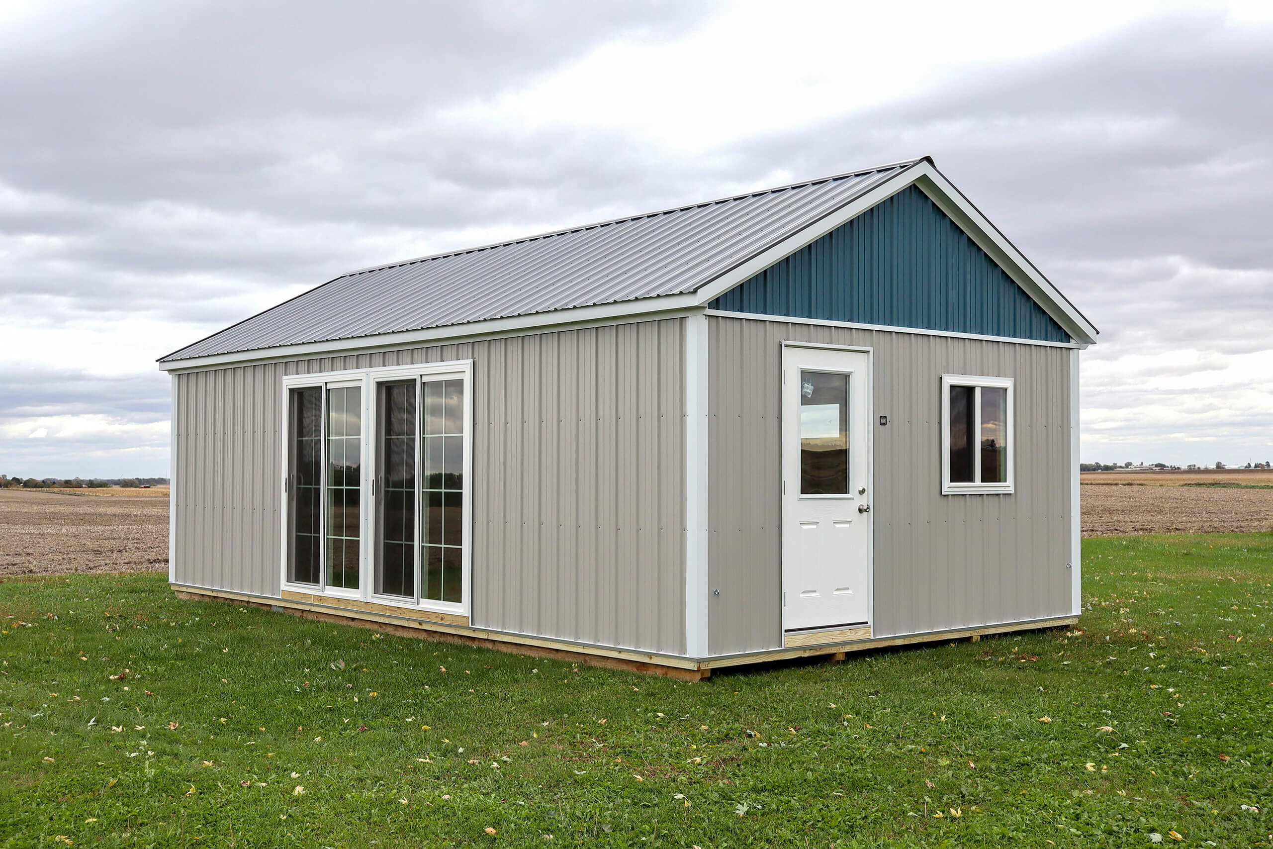 A small, gray and blue metal building with a white door and large glass windows sits on a grassy field under a cloudy sky.