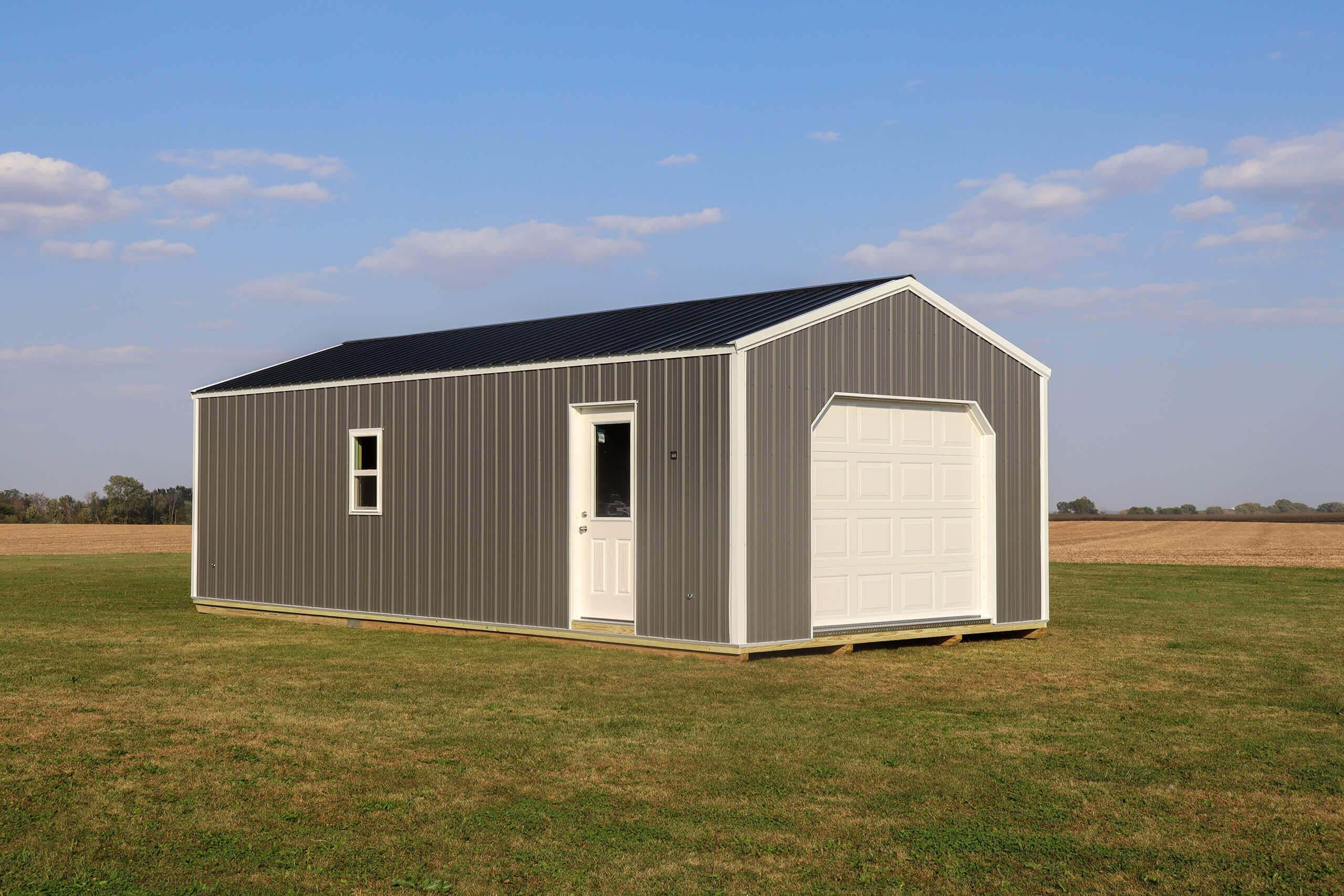 A small gray metal shed with a white door and garage door sits on a grassy field under a partly cloudy sky.