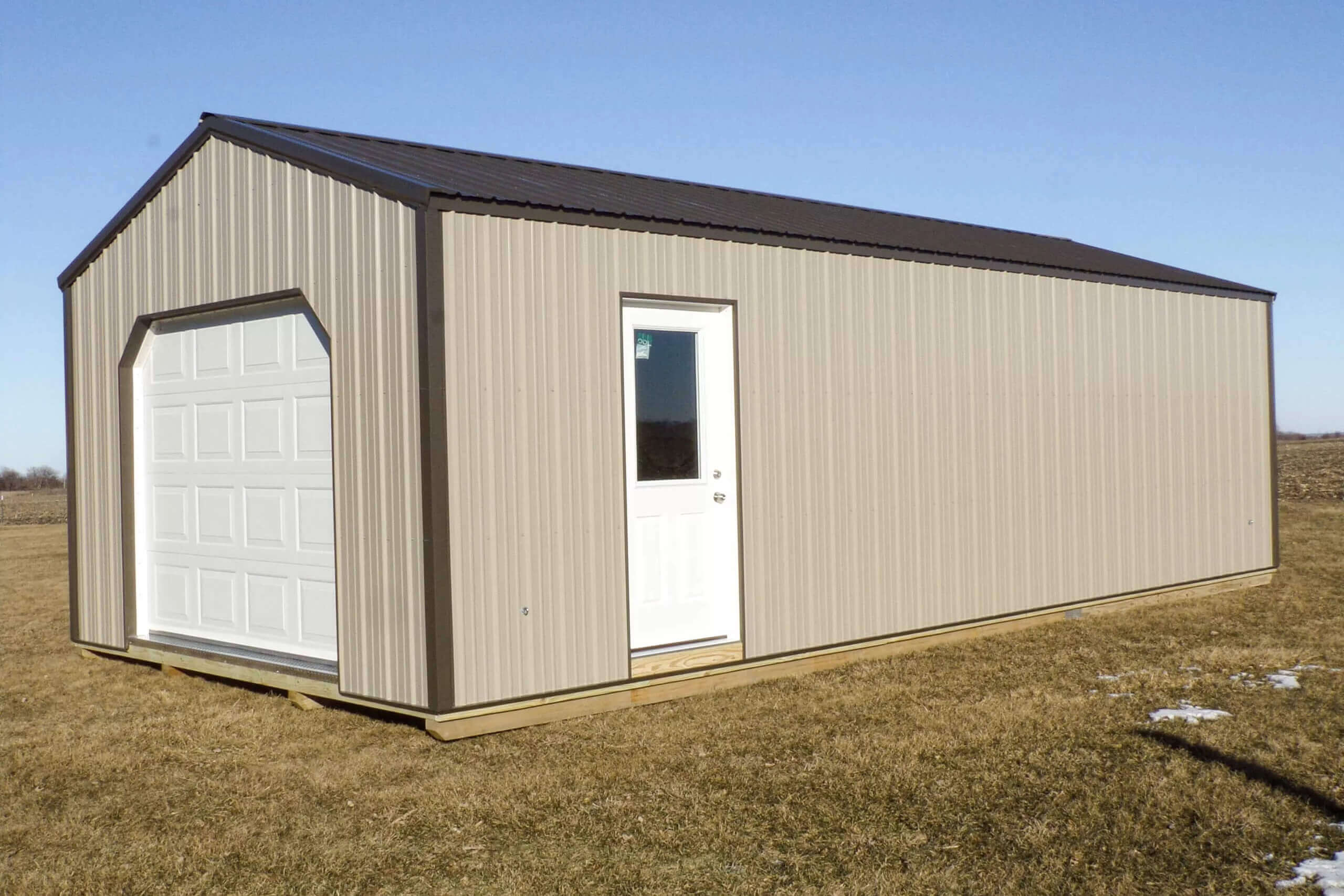 A beige metal shed with a white garage door and a white entry door, standing on a grassy field under a clear blue sky.
