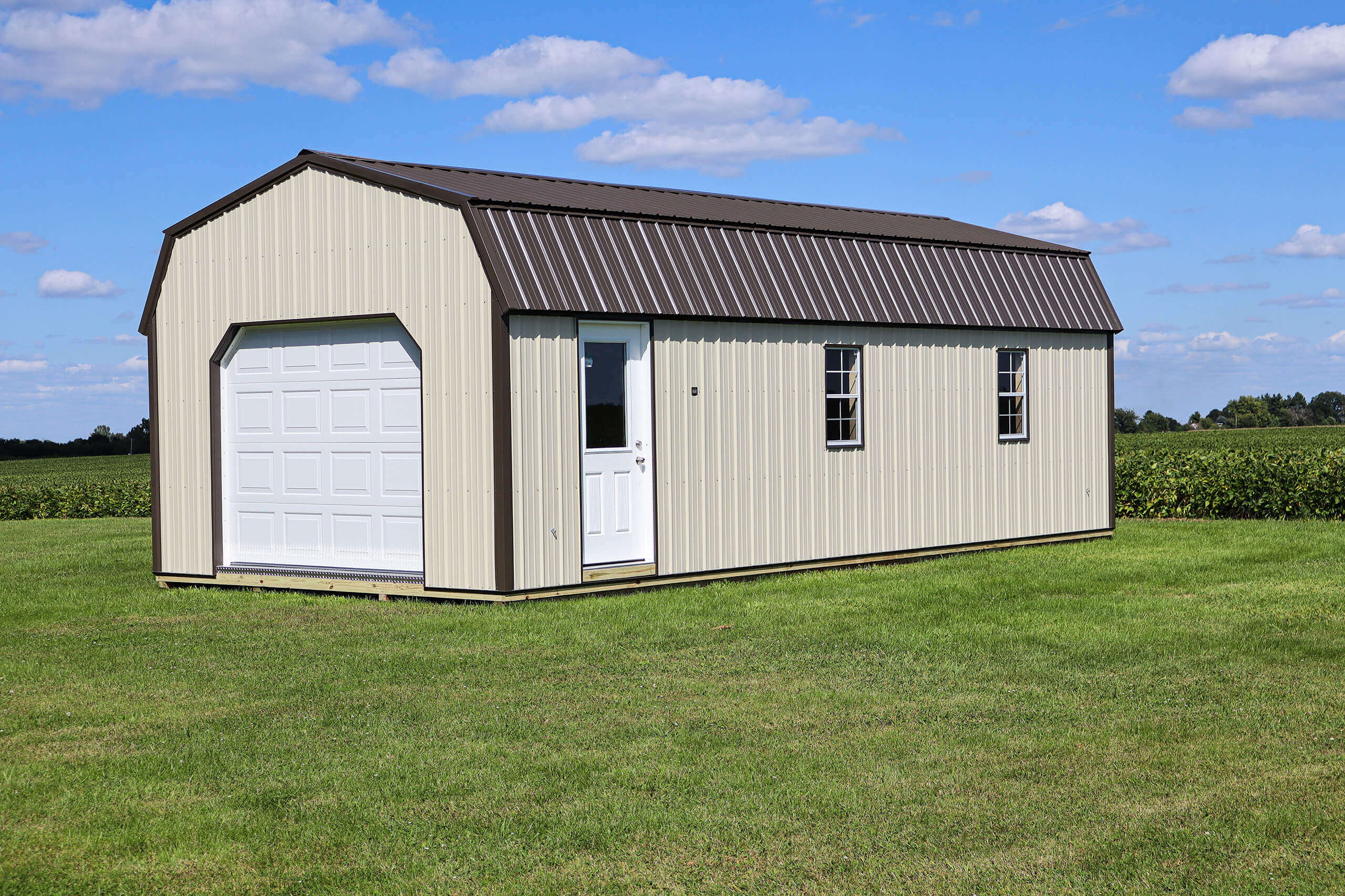 A beige metal shed with a brown roof, white garage door, entry door, and two windows sits on a grassy field under a blue sky with scattered clouds.