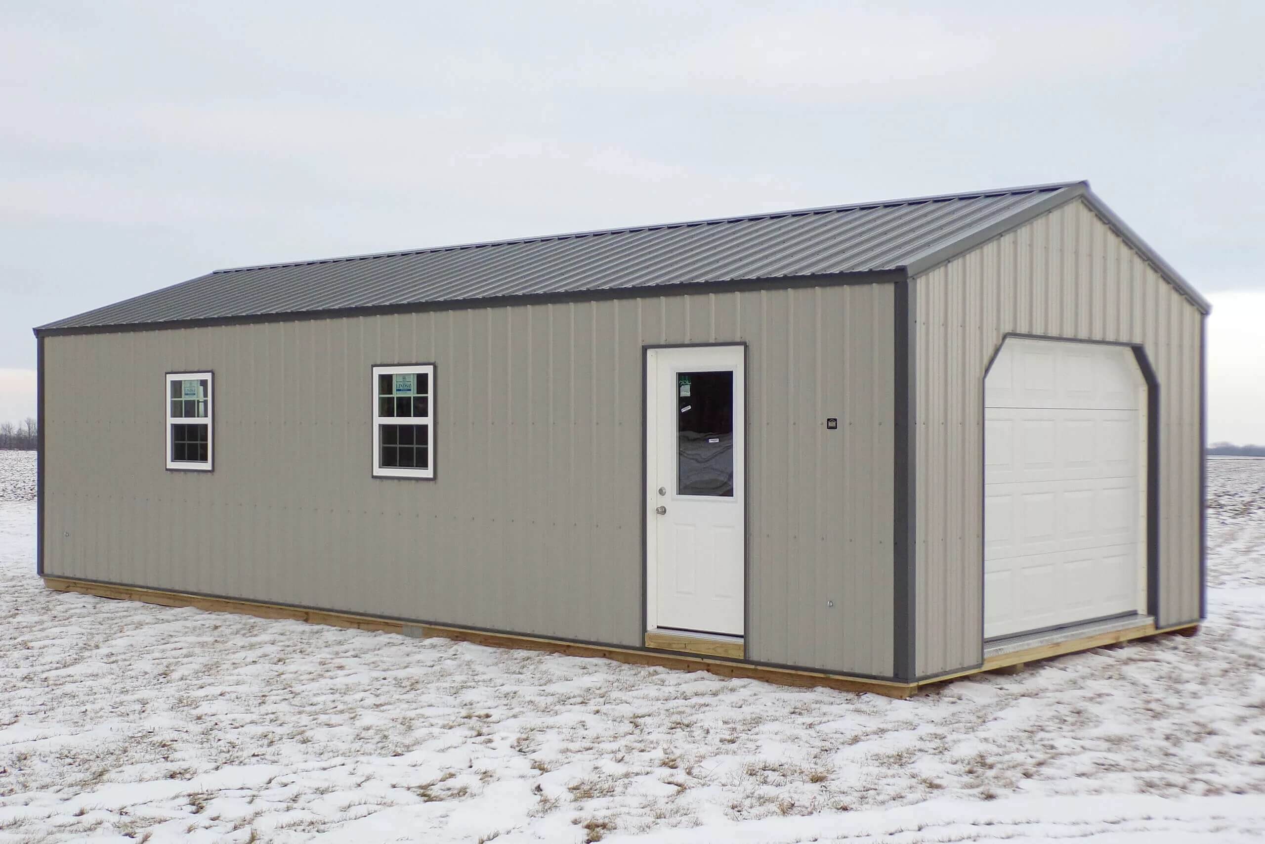 Gray metal shed with two windows, a white entry door, and a garage door, situated on a snowy, flat landscape under an overcast sky.
