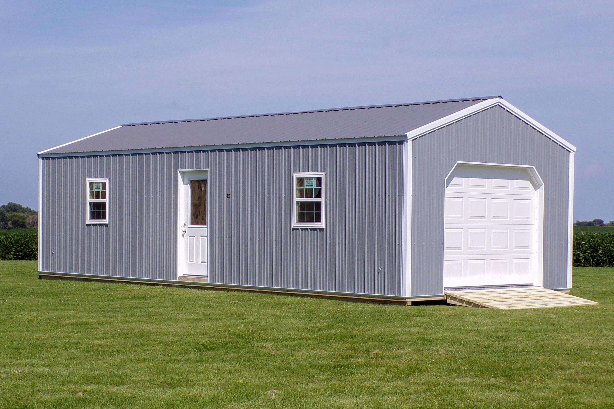 Gray metal shed with a white door, two windows, and a garage door, standing on a grassy field under a clear sky.