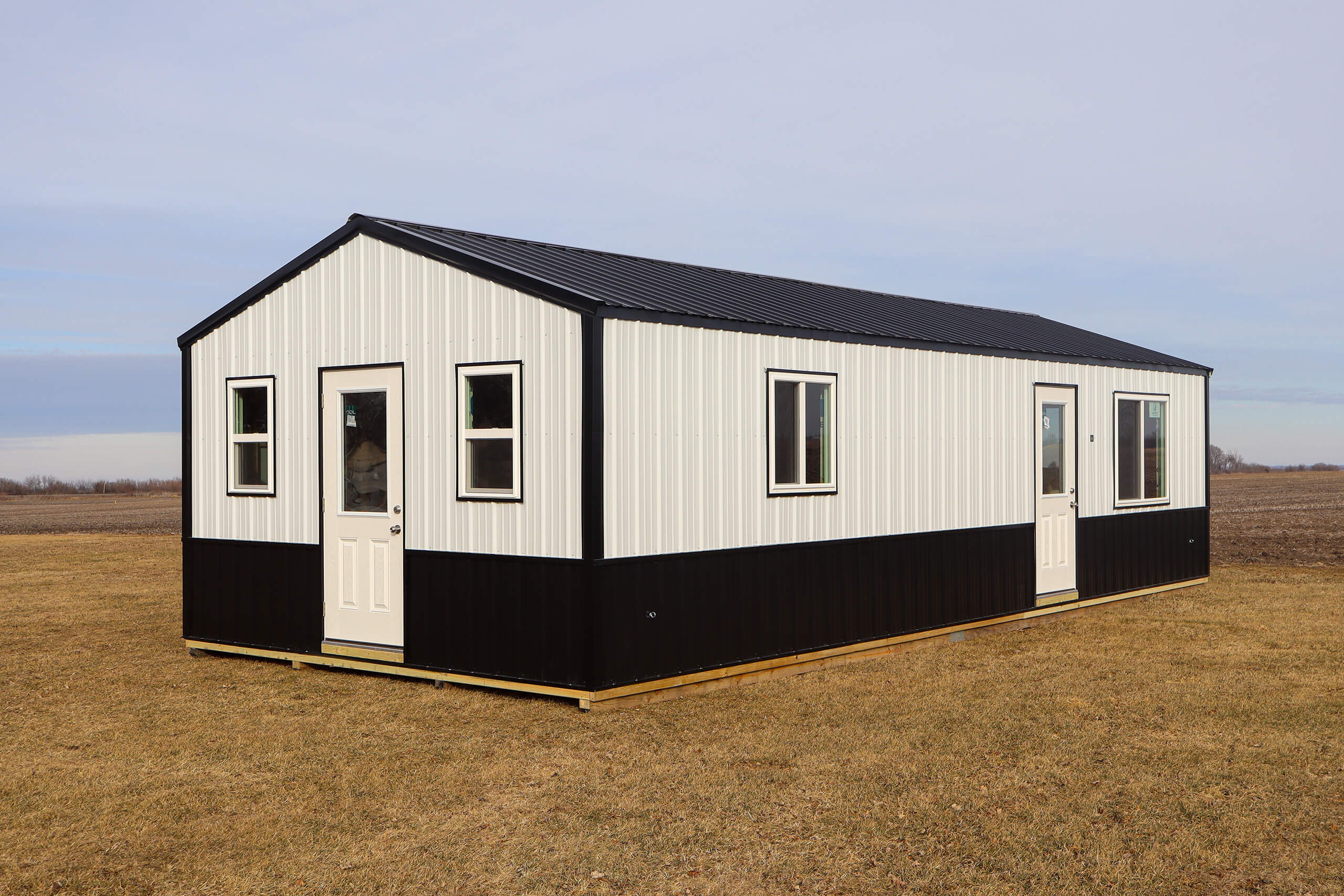 A small, rectangular, black and white metal building with two doors and several windows sits on a grassy field under a clear sky.