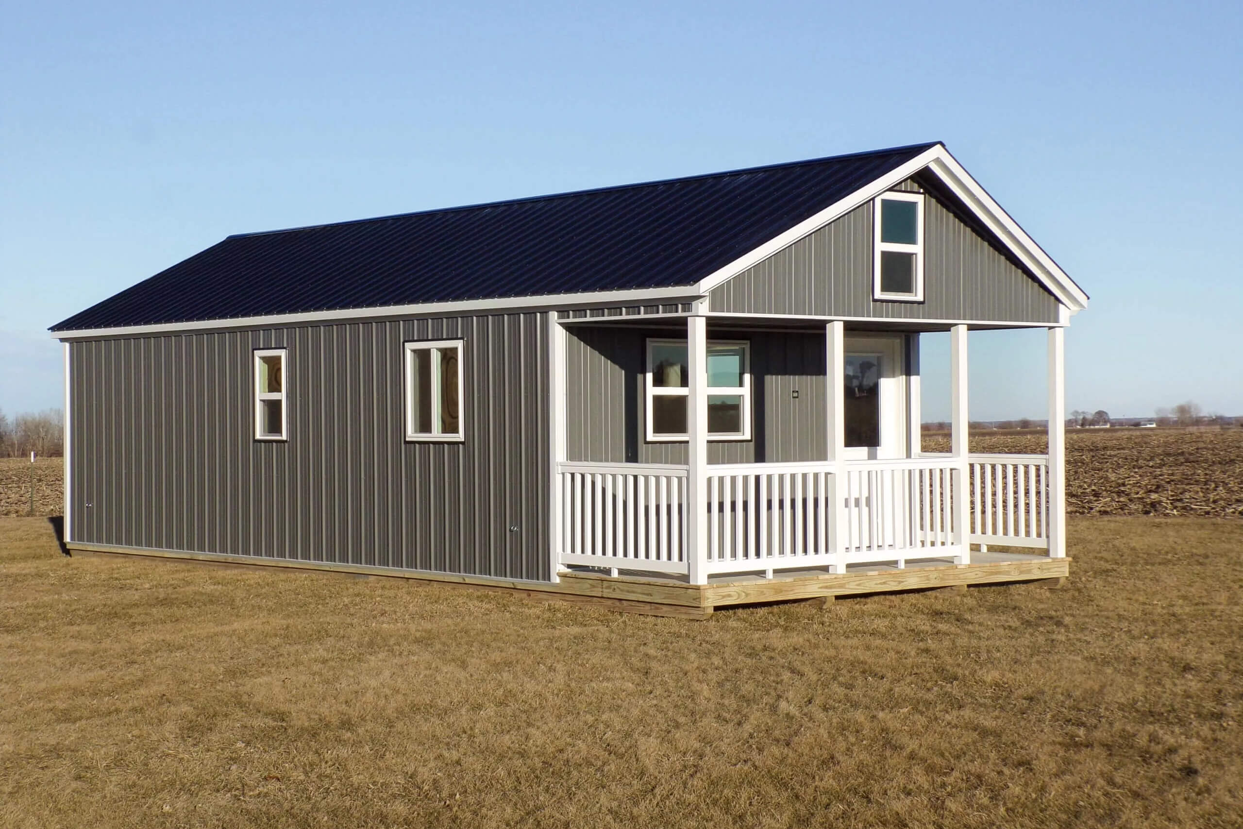 A small gray house with a black roof, white trim, and front porch sits on a grassy field under a clear blue sky.