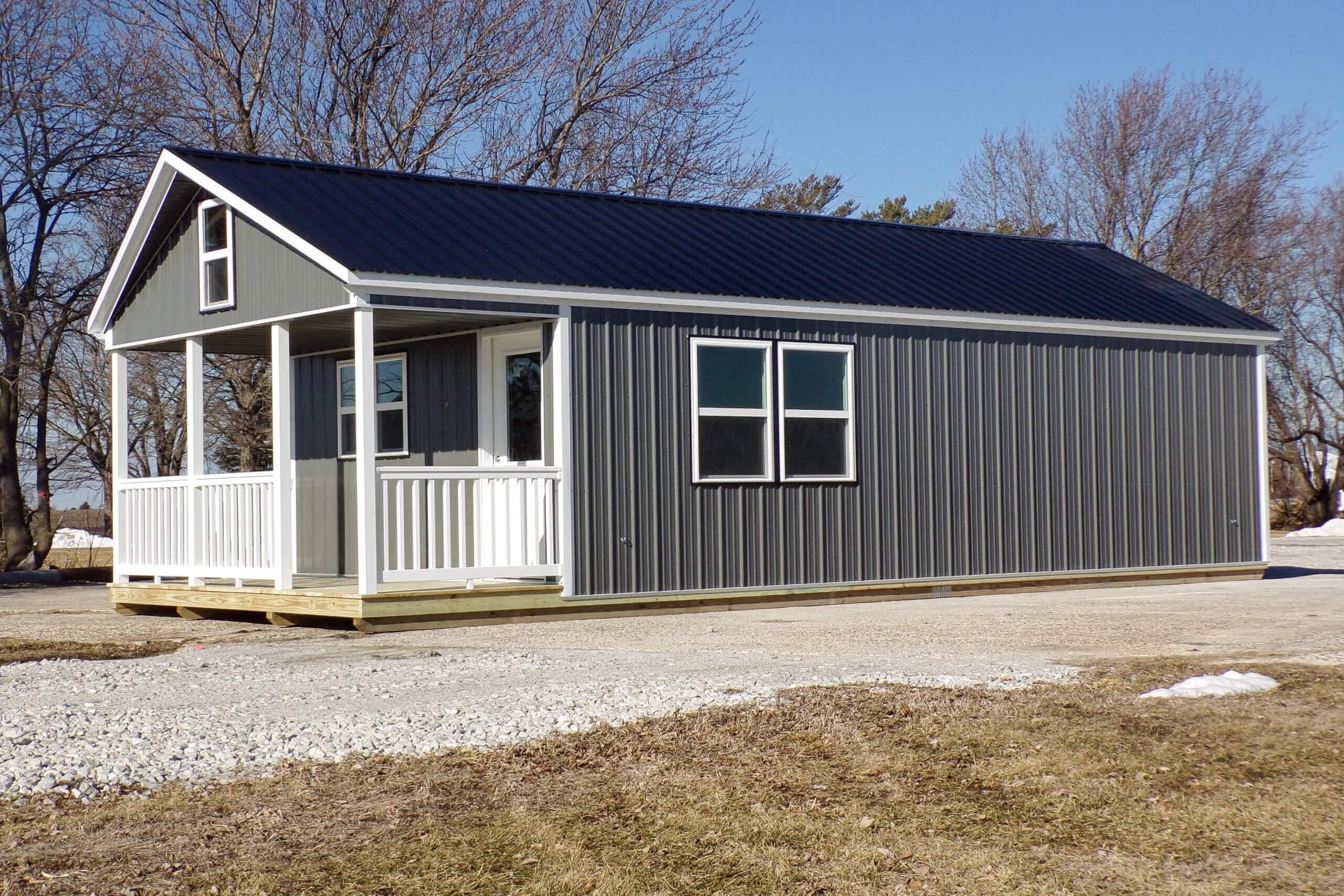 Small gray metal building with a front porch, white trim, and black roof, standing on a gravel driveway with leafless trees in the background.