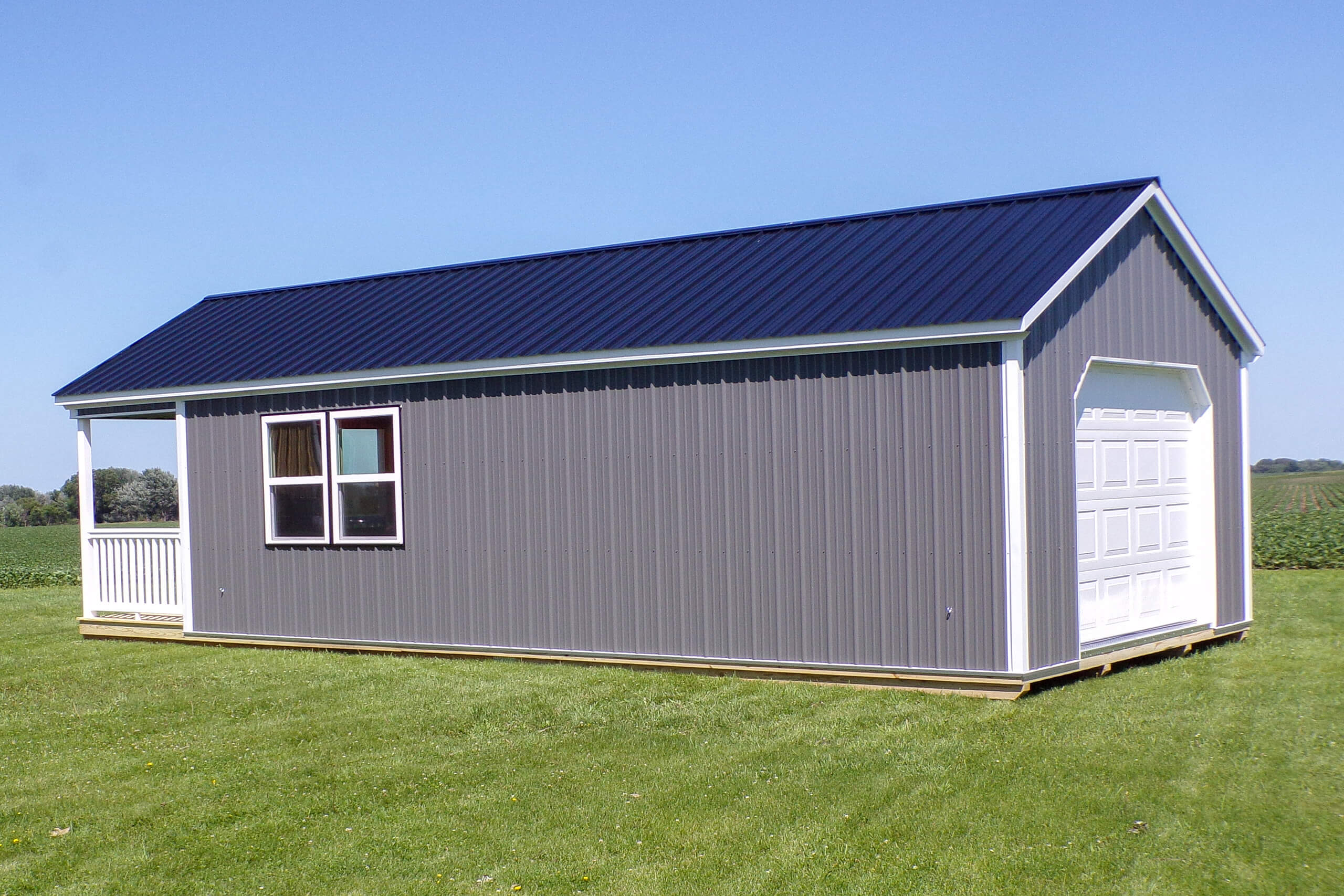 Gray metal shed with a black roof, two windows, a white garage door, and a small porch, situated on a grassy field under a clear blue sky.