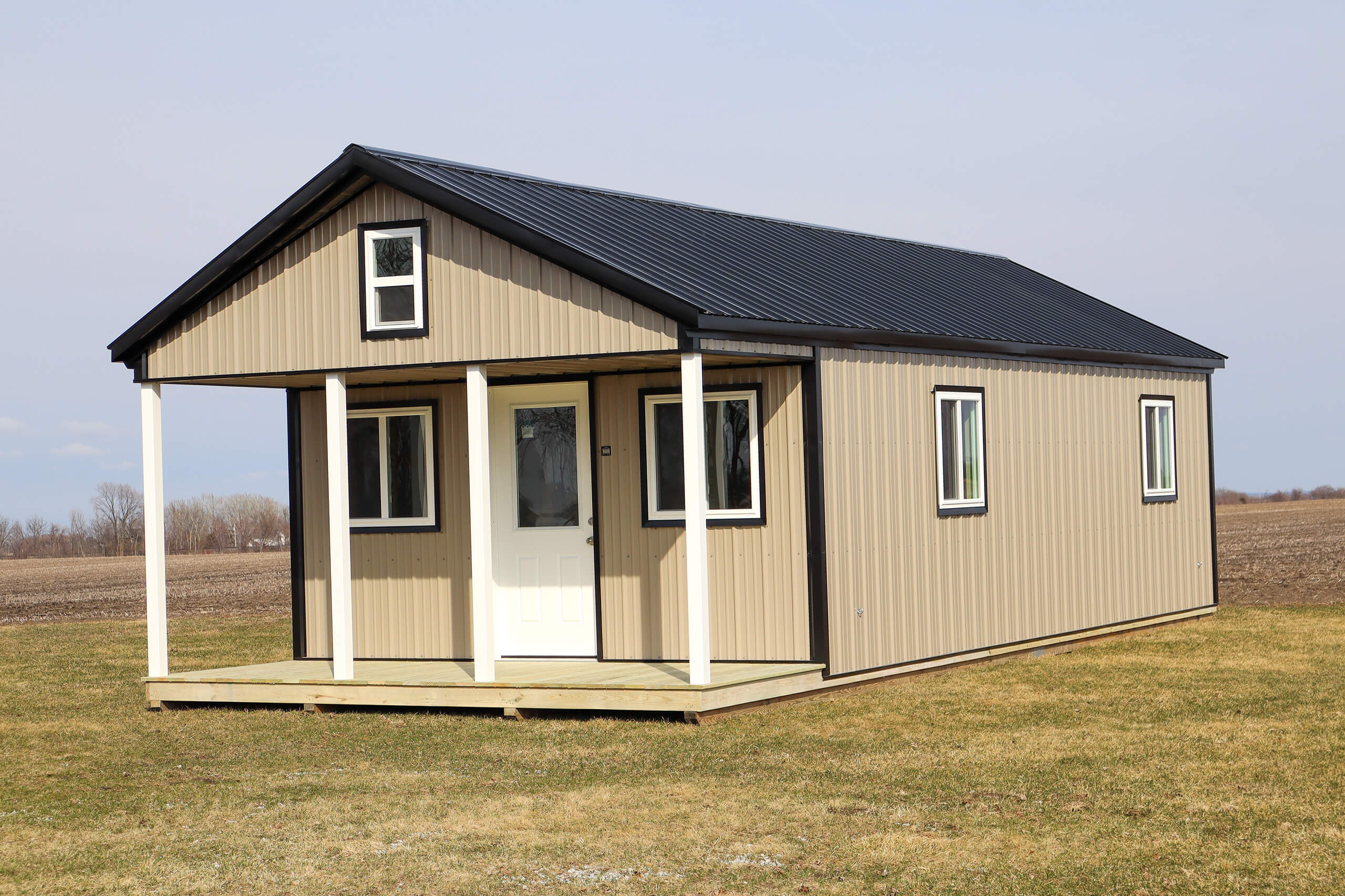 A small beige and black metal cabin with a front porch and white door, standing on a grassy area beside a field under a clear sky.