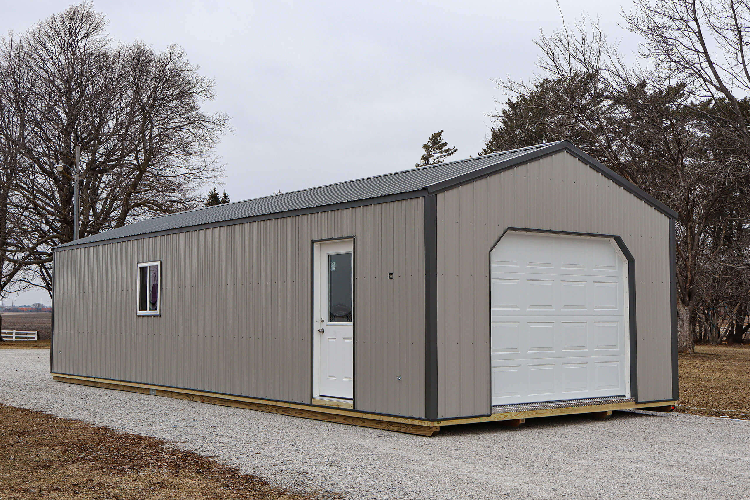A prefabricated metal building with a white door, window, and garage door sits on a gravel driveway, surrounded by bare trees.