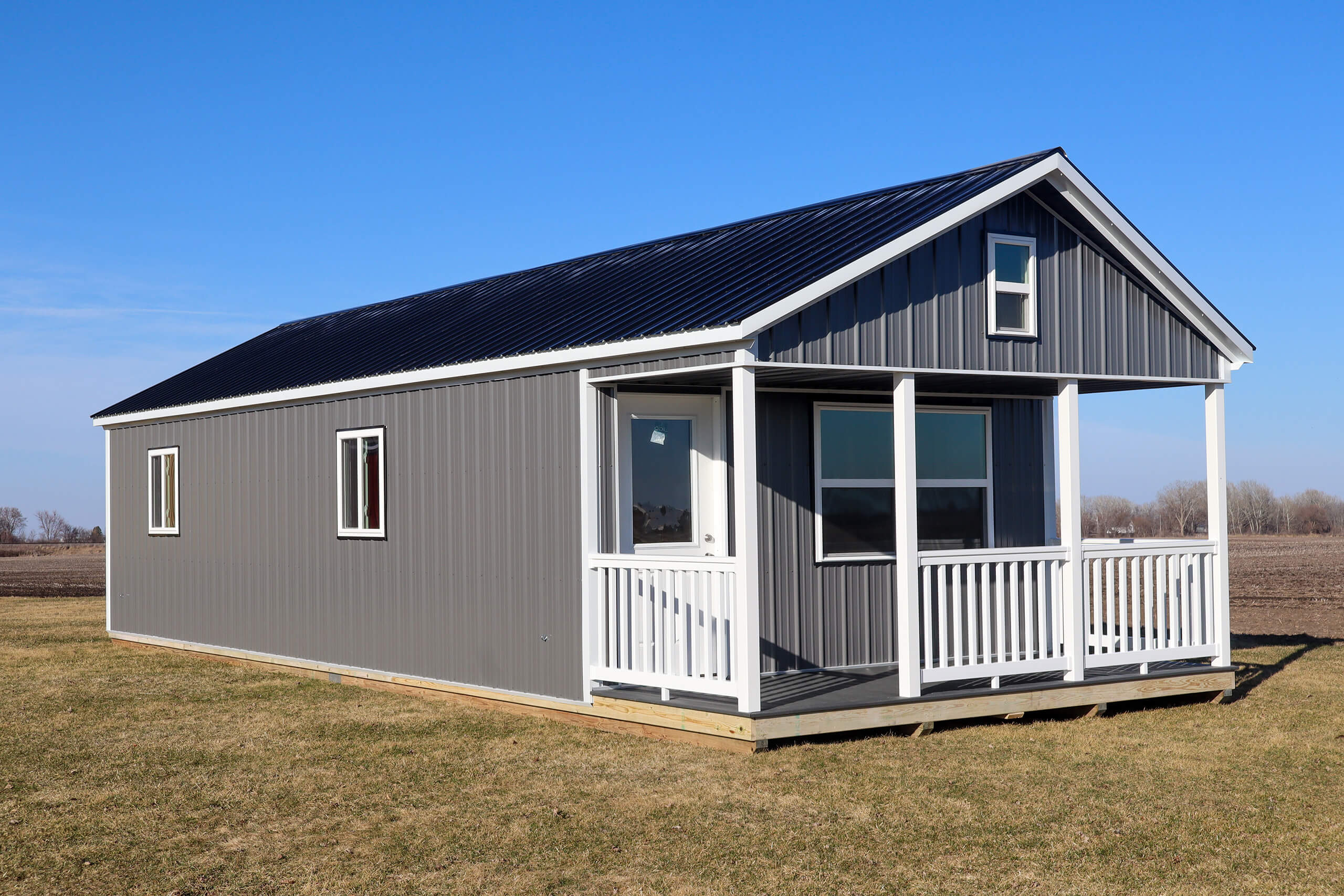 A small gray cabin with a black metal roof, white trim, and a front porch sits on a grassy field under a clear blue sky.