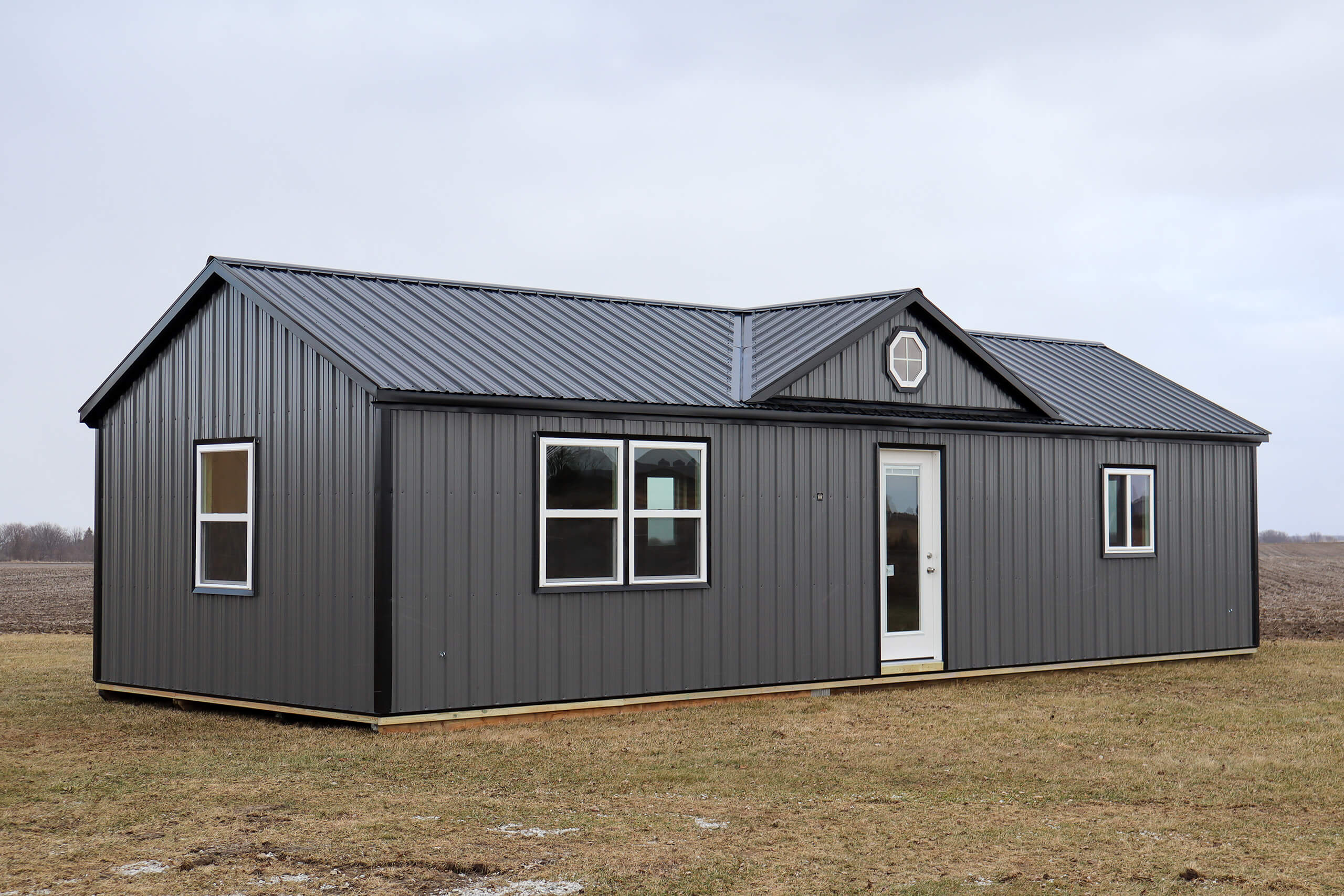 A small, gray metal building with a gable roof, white-framed windows, and a single glass door stands on a grassy field under an overcast sky.