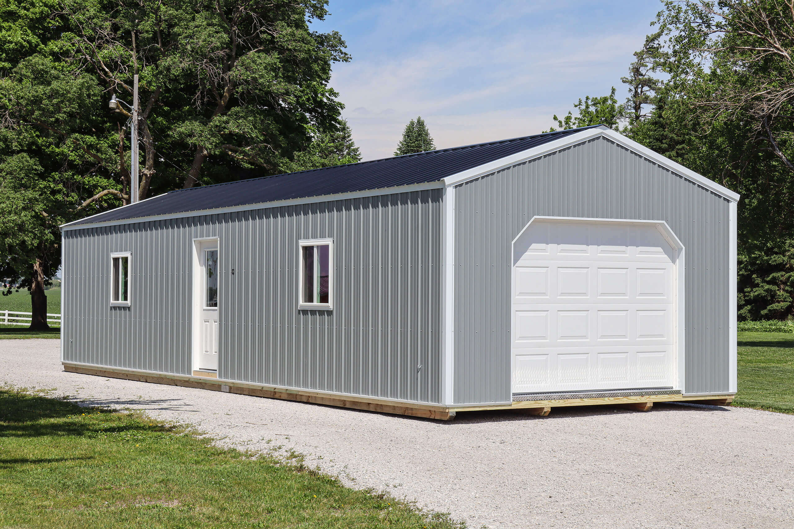 A gray metal garage building with a white entry door, two windows, and a white roll-up garage door sits on a gravel driveway next to a grassy area and trees.
