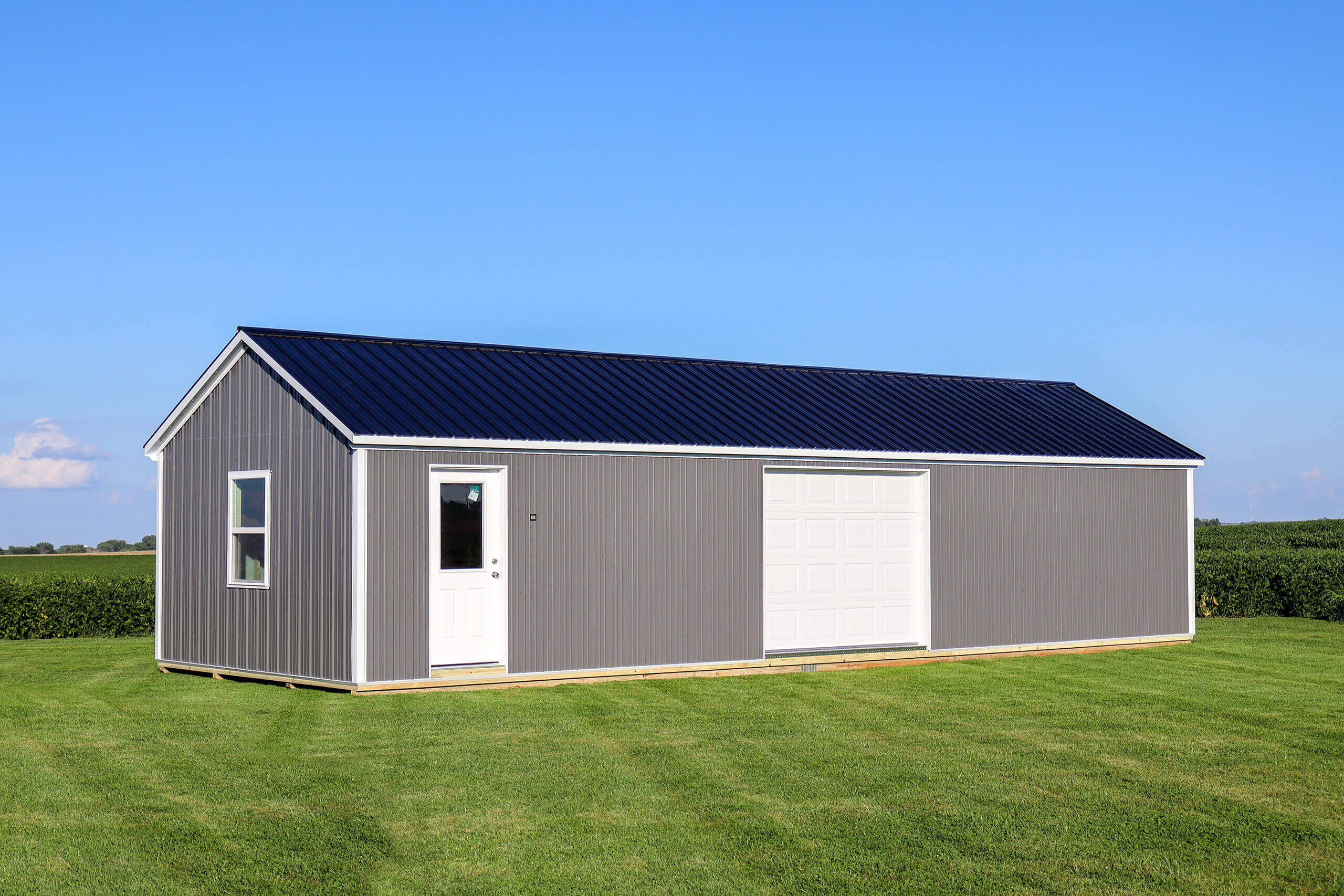 Gray metal storage shed with a white door, window, and large garage door, sits on green grass under a clear blue sky.