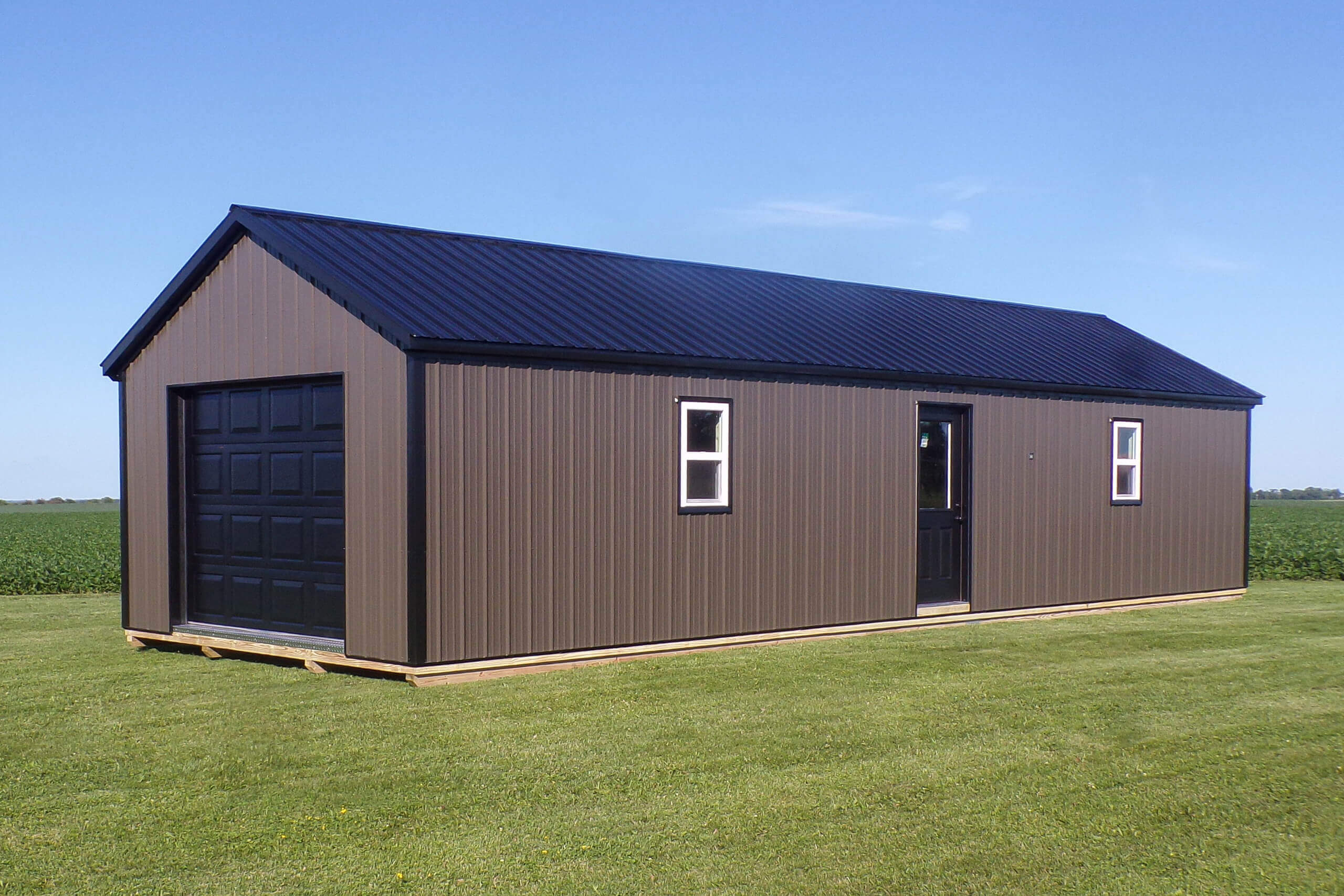 A brown metal shed with a black garage door, two windows, and a single entry door stands on a grassy field under a clear blue sky.