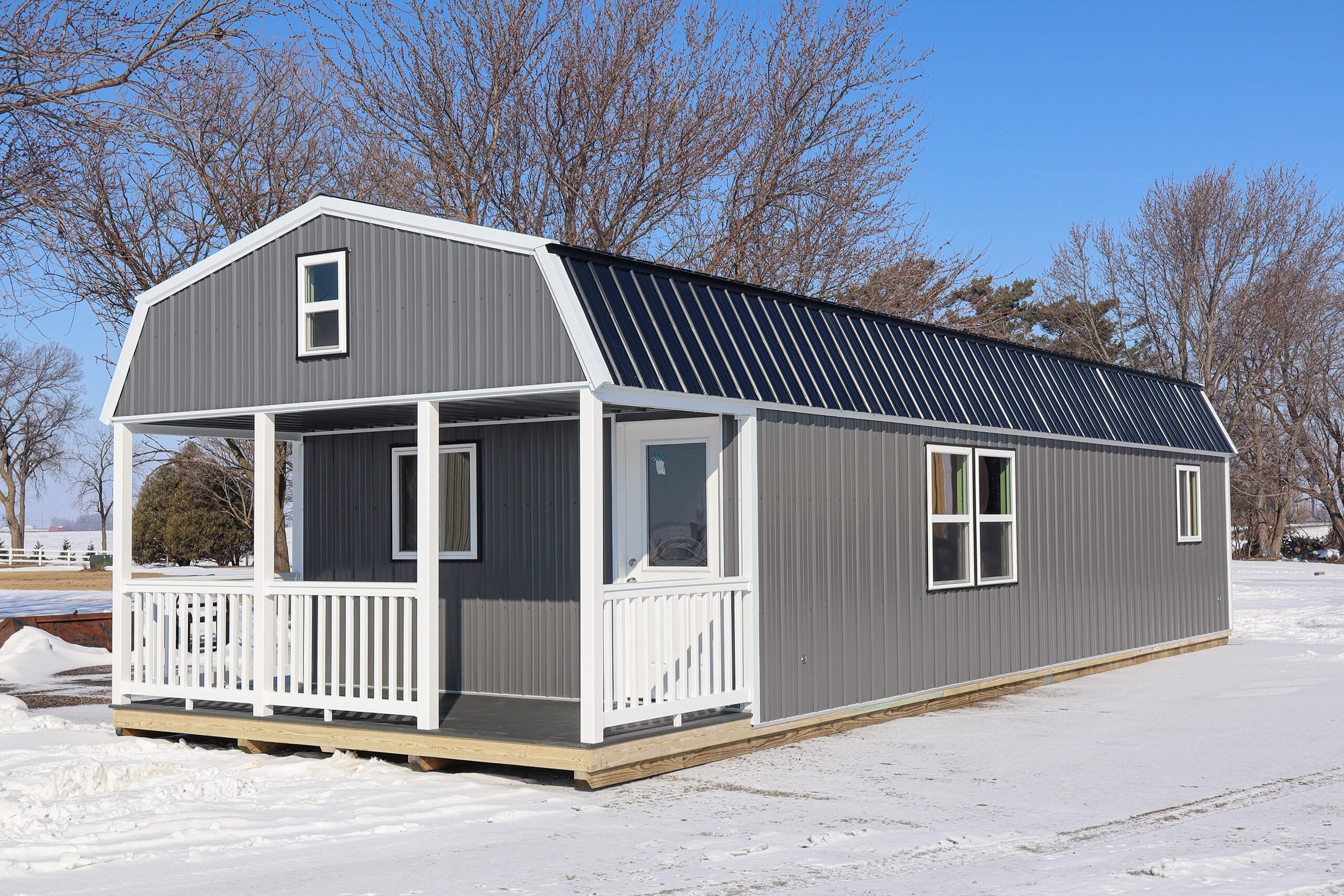 A small gray metal-sided house with a black roof and a white porch sits on a snowy lot under a clear blue sky.