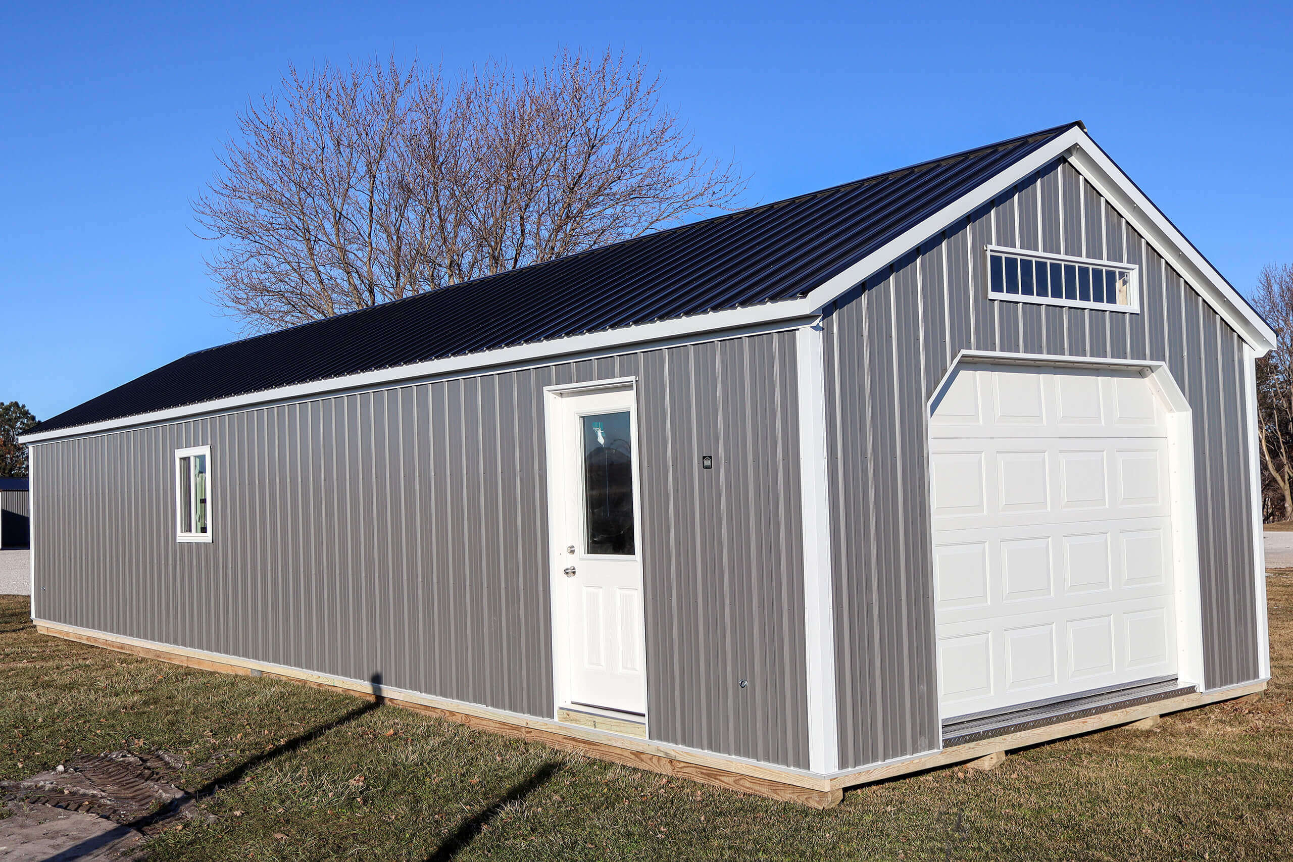 A small gray metal building with a white garage door, side entry door, and a window, sits on grass under a clear blue sky.