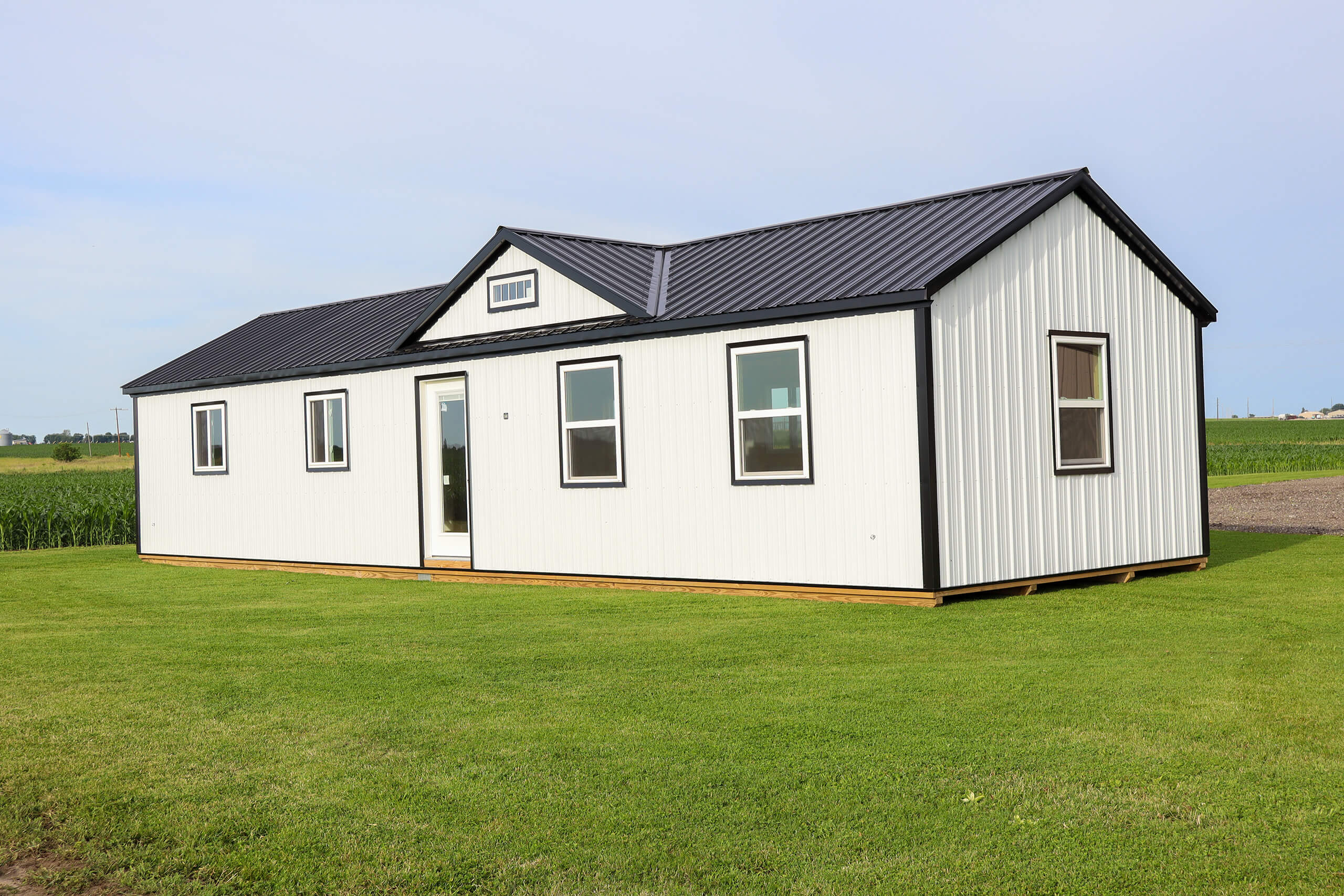 A small white modular home with a black metal roof sits on a grassy lawn, with fields and a clear sky in the background.