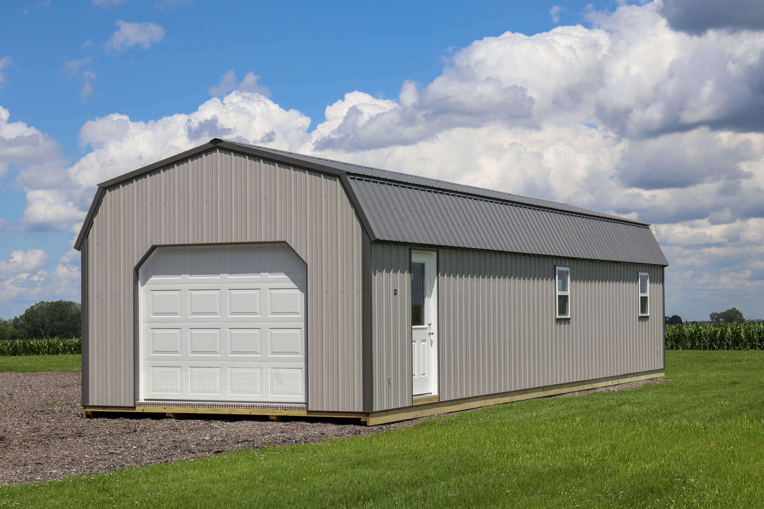 A gray metal storage shed with a white garage door and side entry door stands on a grassy area under a partly cloudy sky.