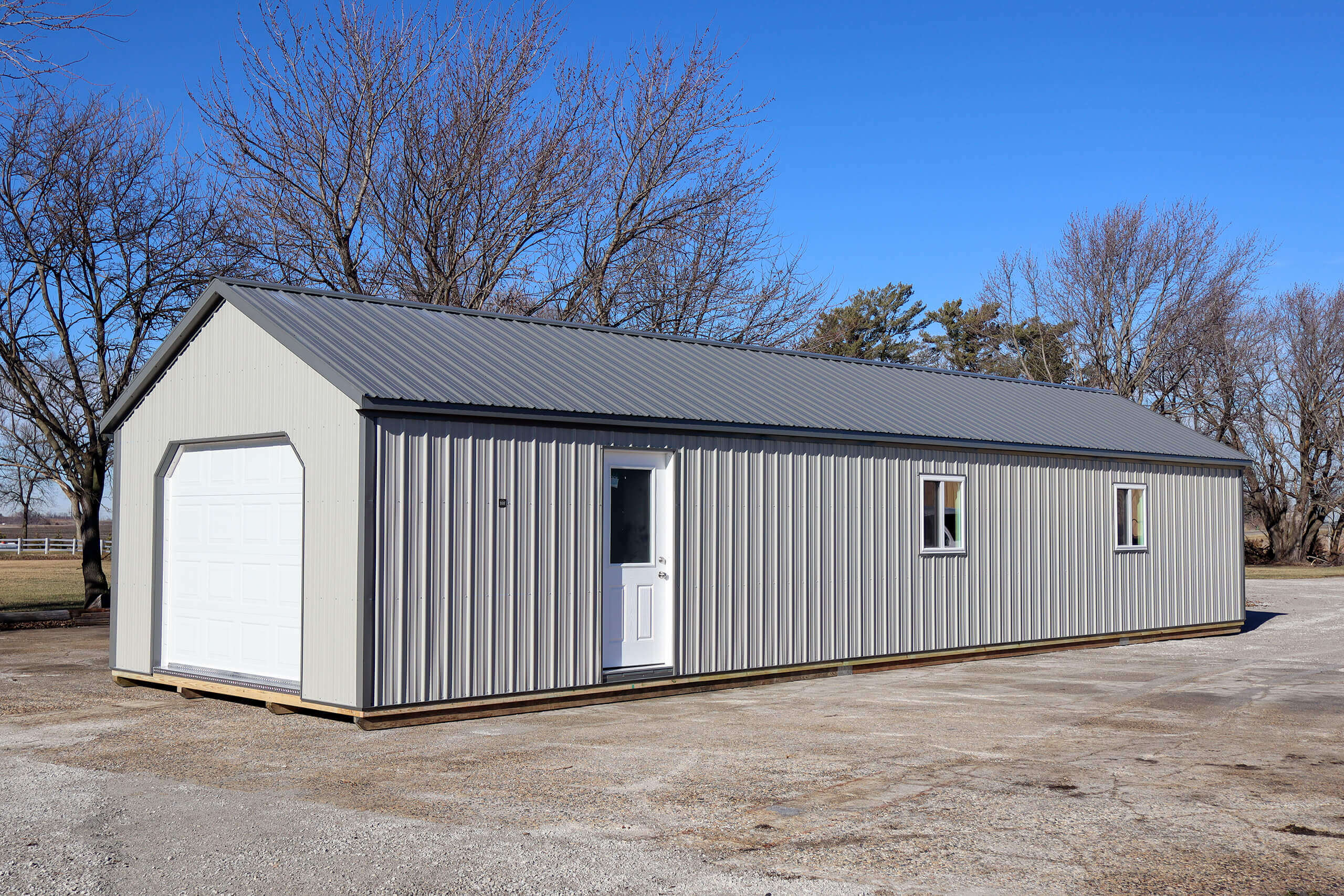 A gray metal shed with a white garage door, a white entry door, and two small windows, set on a gravel lot with leafless trees in the background under a clear blue sky.