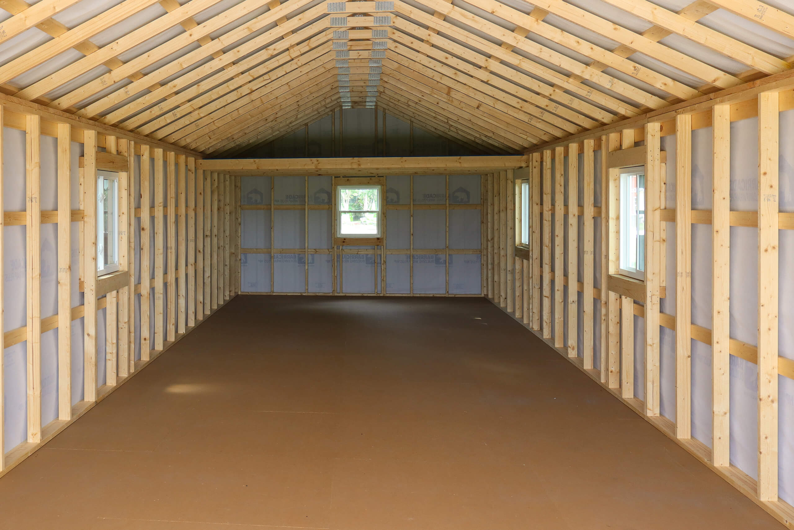 Interior of an unfinished wooden building with exposed framing, windows on both sides, and a door with a window at the far end. Floor appears to be plywood.