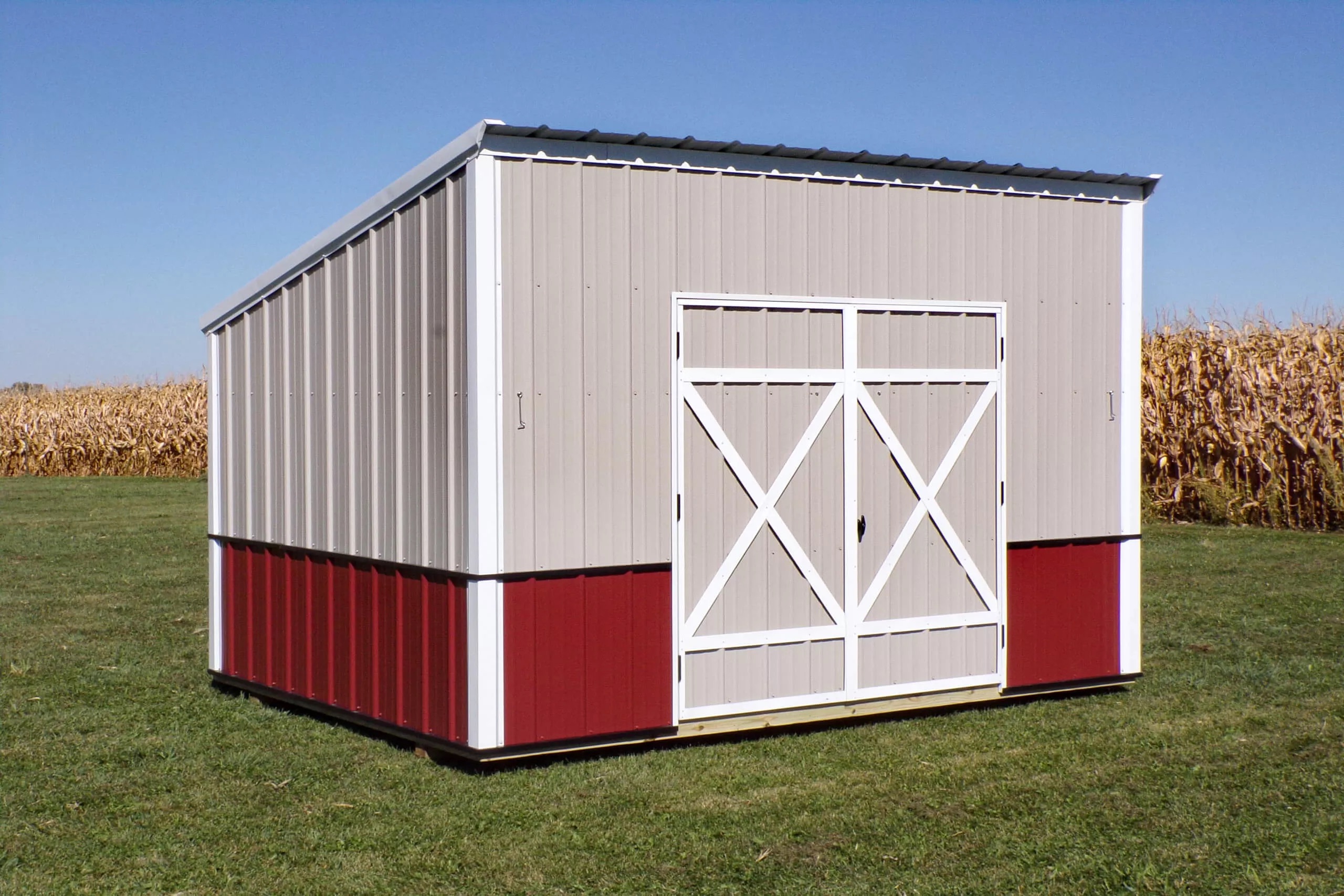 A small metal shed with beige and red siding and white double doors stands on grass near a cornfield under a clear blue sky.