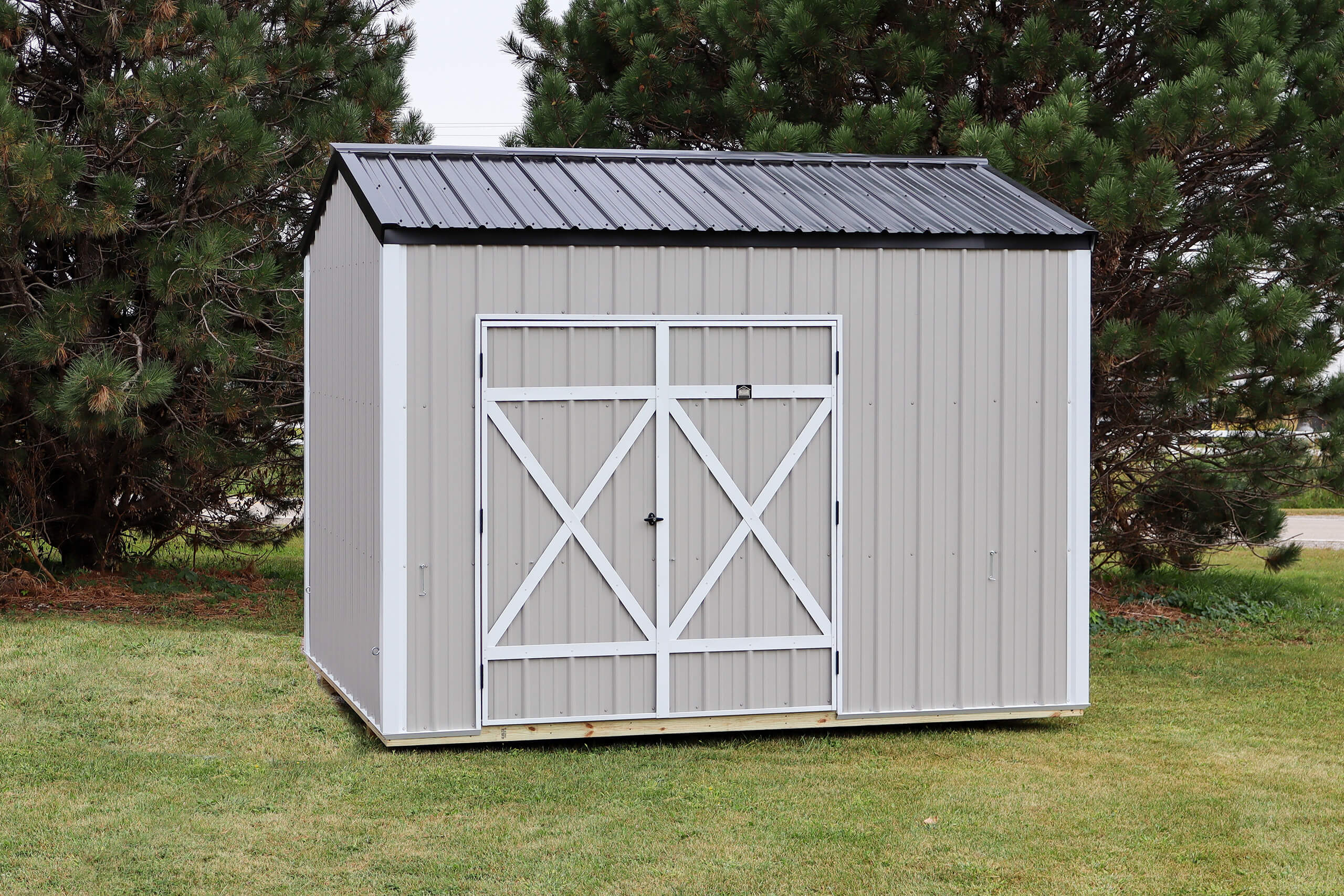 A gray metal storage shed with a black roof and white double doors stands on a grassy lawn, surrounded by pine trees.