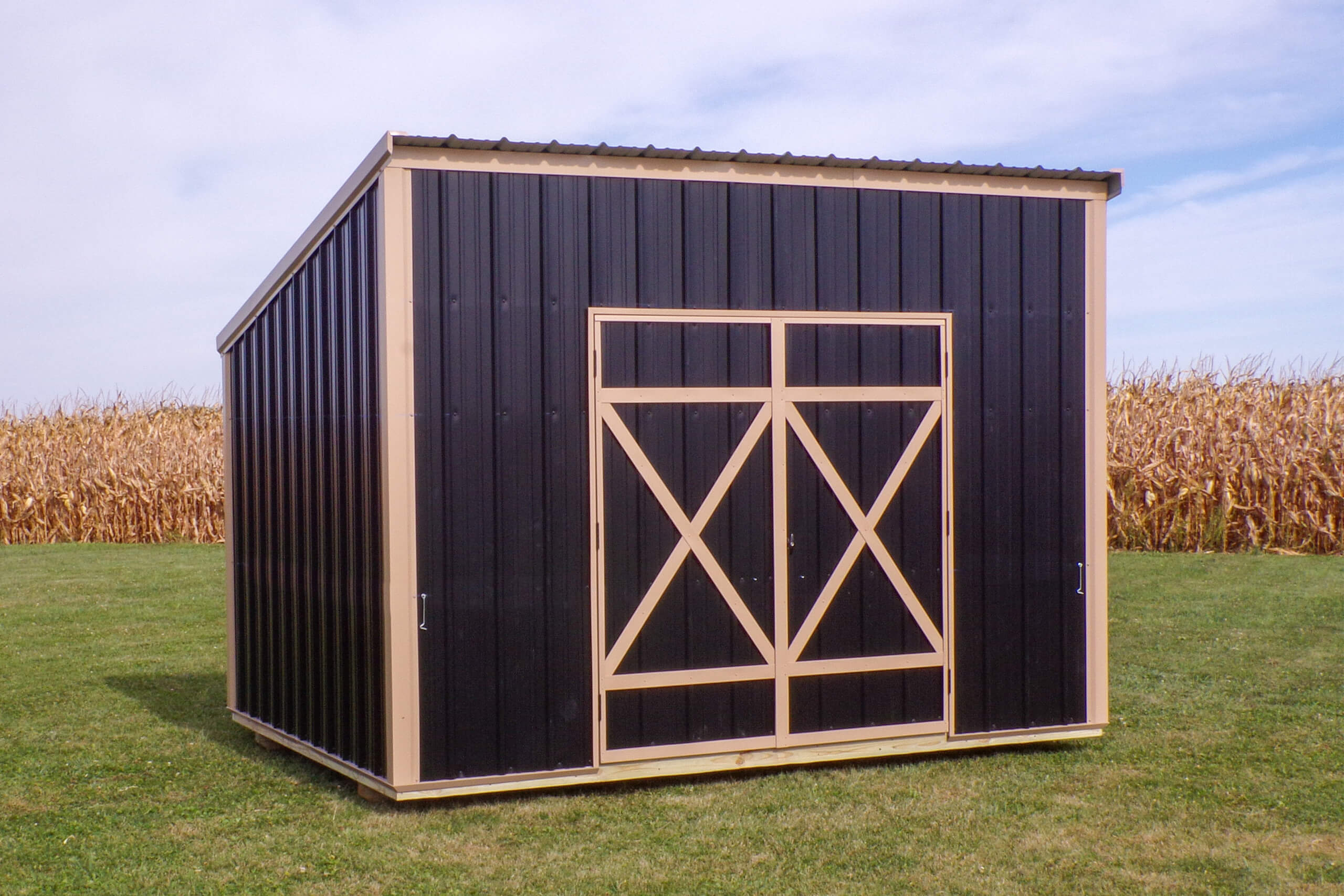 A small black metal shed with tan trim stands on grass near a cornfield under a partly cloudy sky.