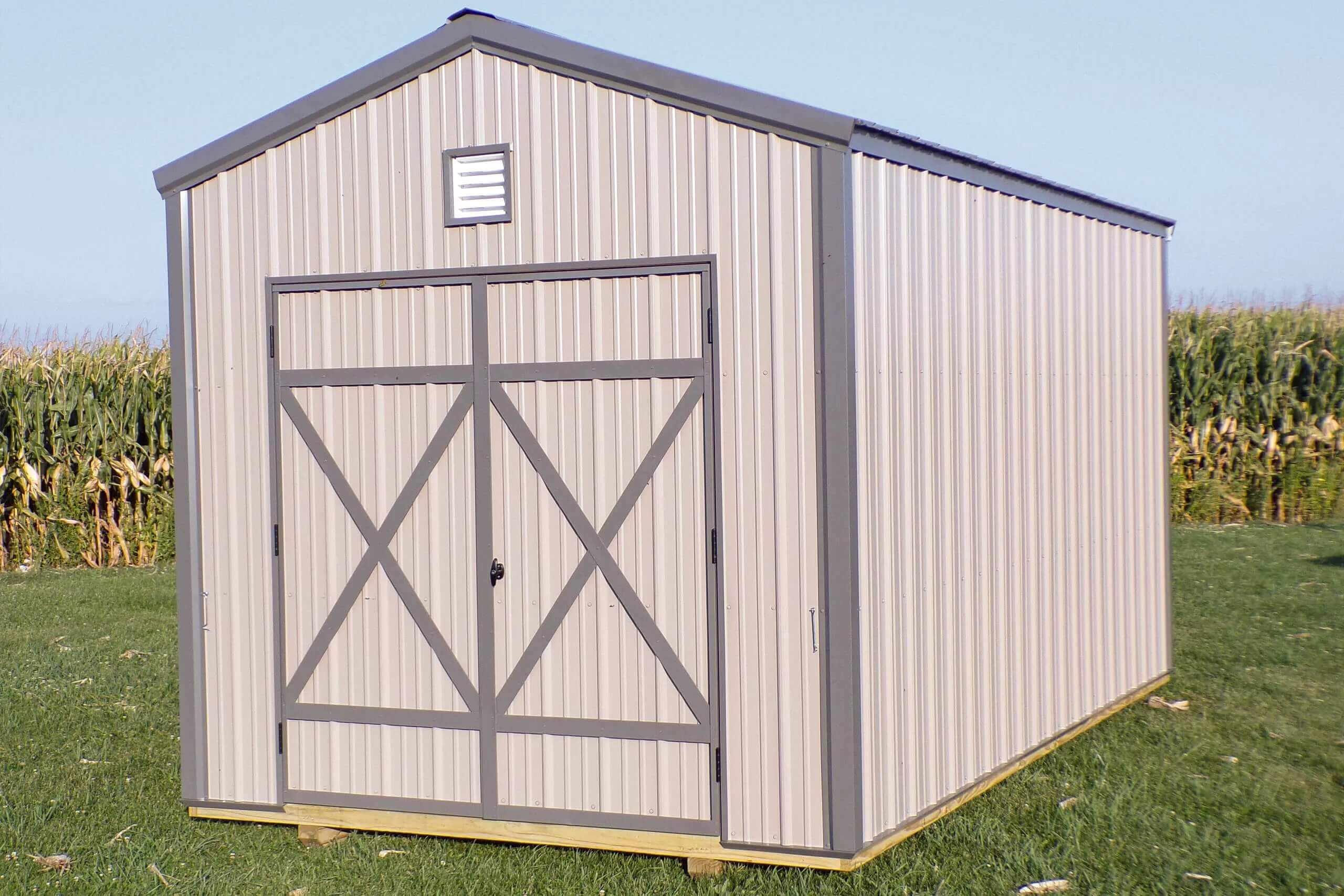 A beige and gray metal storage shed with double doors stands on a grassy area, with a cornfield visible in the background.