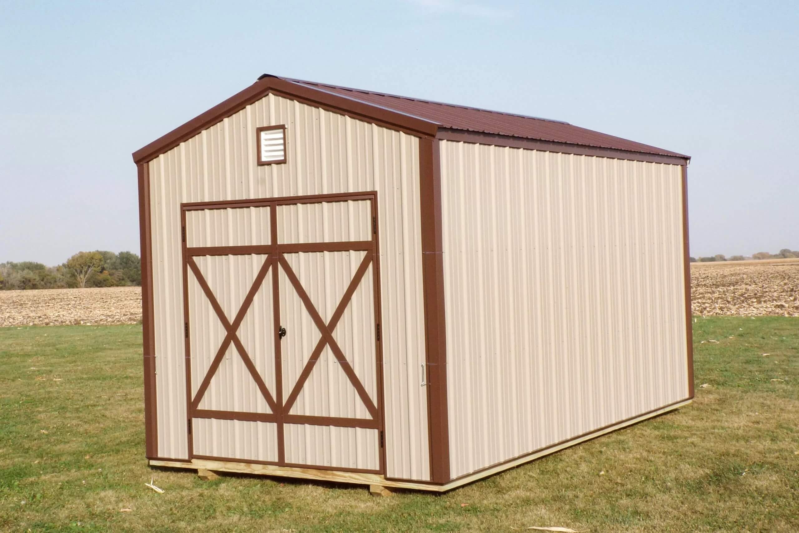 A beige and brown metal storage shed with double doors stands on a grassy field under a clear sky.