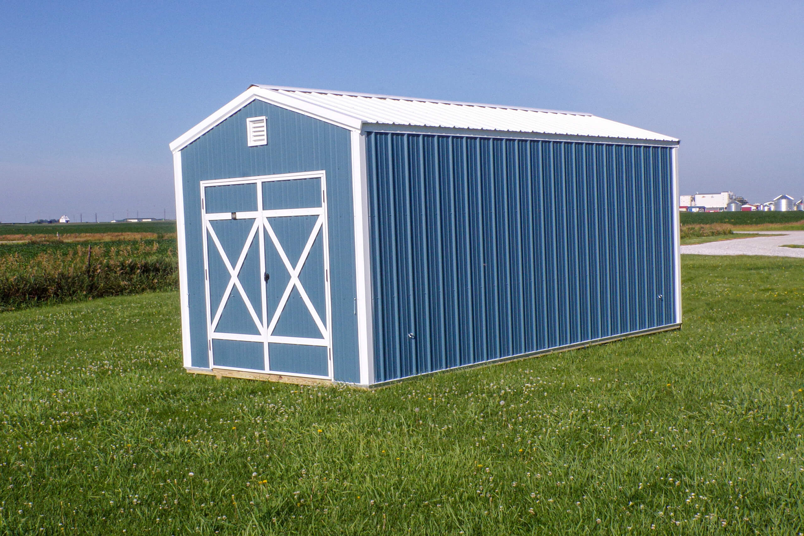 A blue metal storage shed with white trim sits on a grassy field under a clear sky, with some buildings visible in the distant background.
