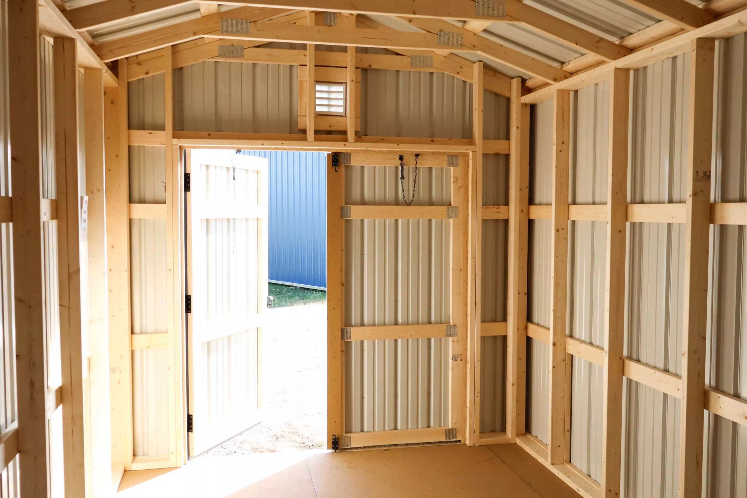 Interior of a small unfinished shed with exposed wooden framing and metal walls, door open to the outside, letting in natural light.