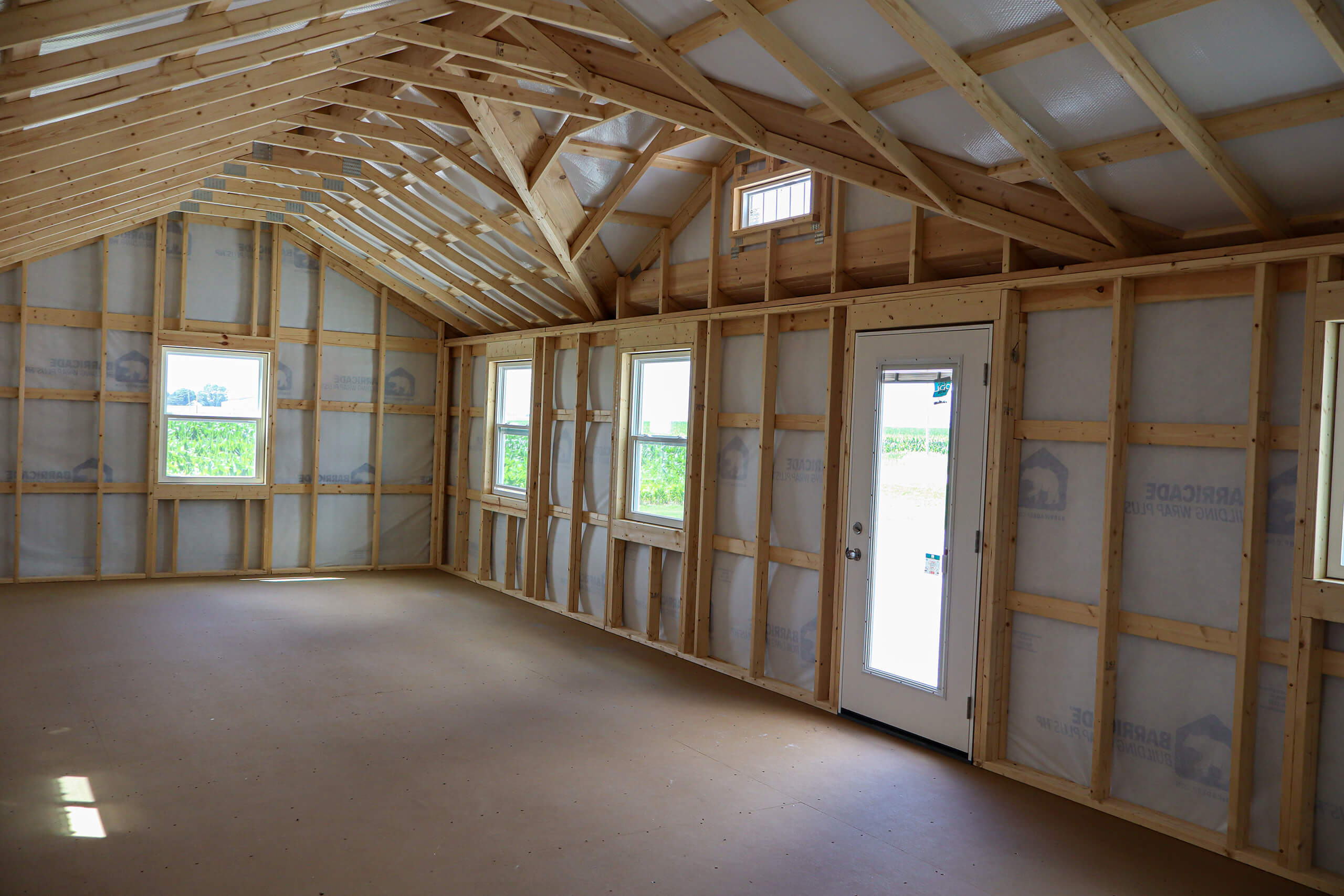 Interior of a partially constructed building showing exposed wooden framing, insulation, three windows, and a door with natural light coming in.