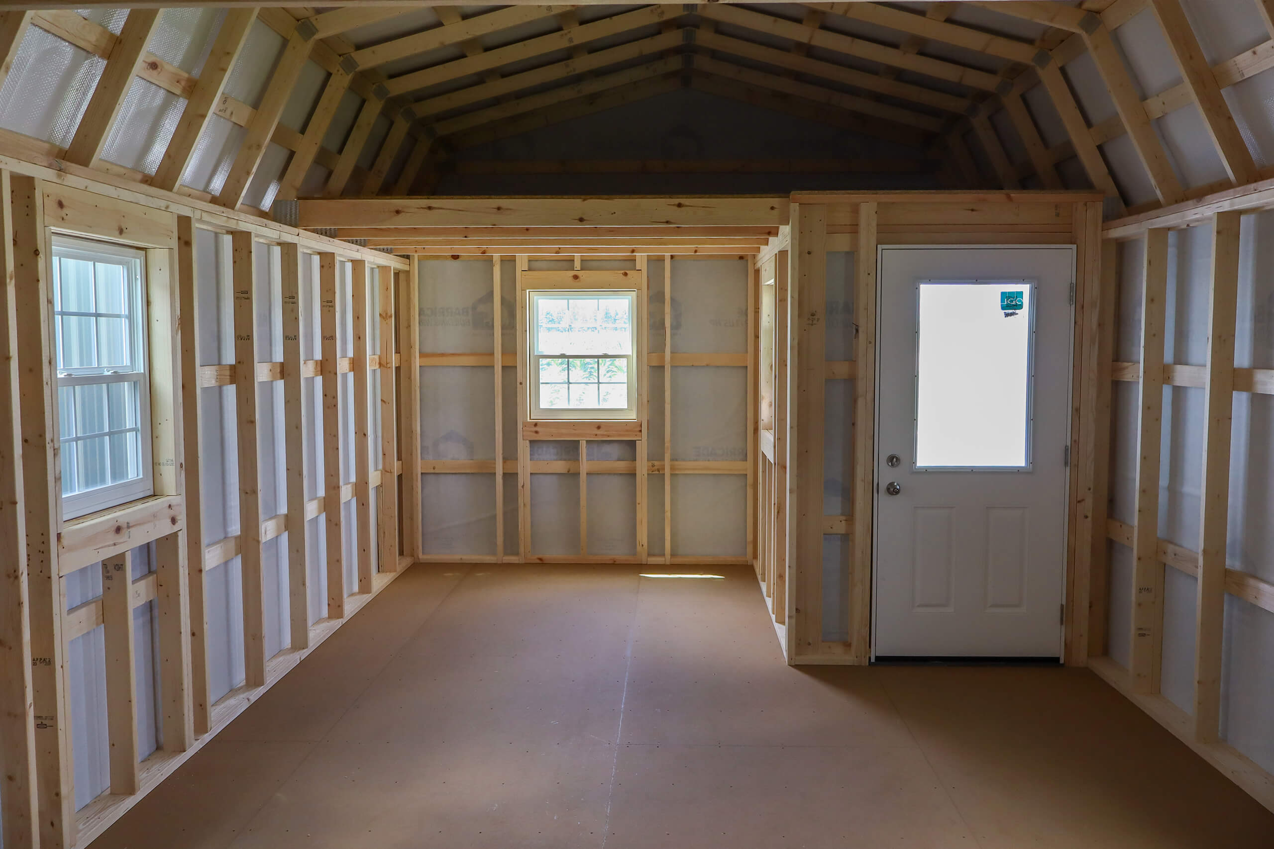 Interior of a small unfinished wooden shed with a door, two windows, exposed framing, and a high ceiling.