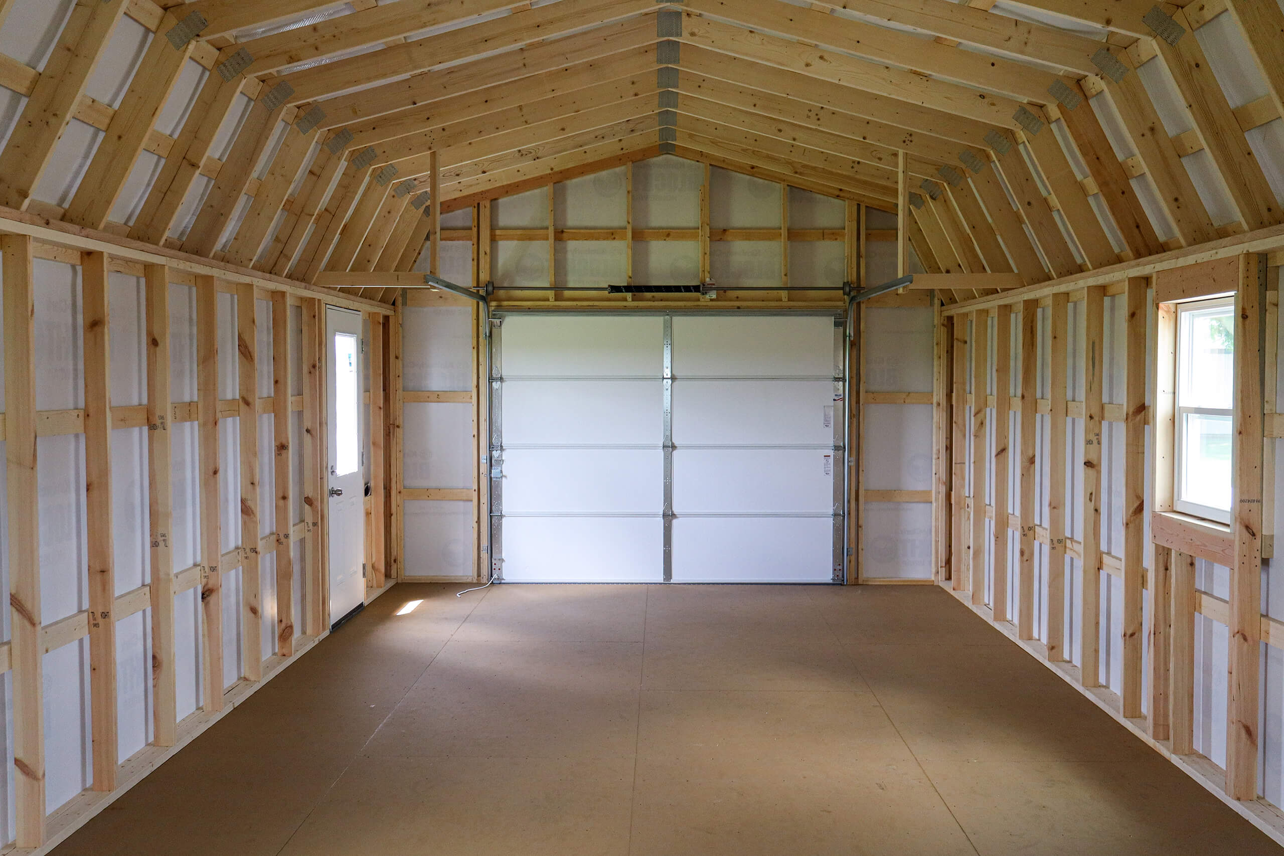 Unfinished interior of a wooden garage or shed with exposed framing, a closed white garage door, windows on both sides, and a bare plywood floor.
