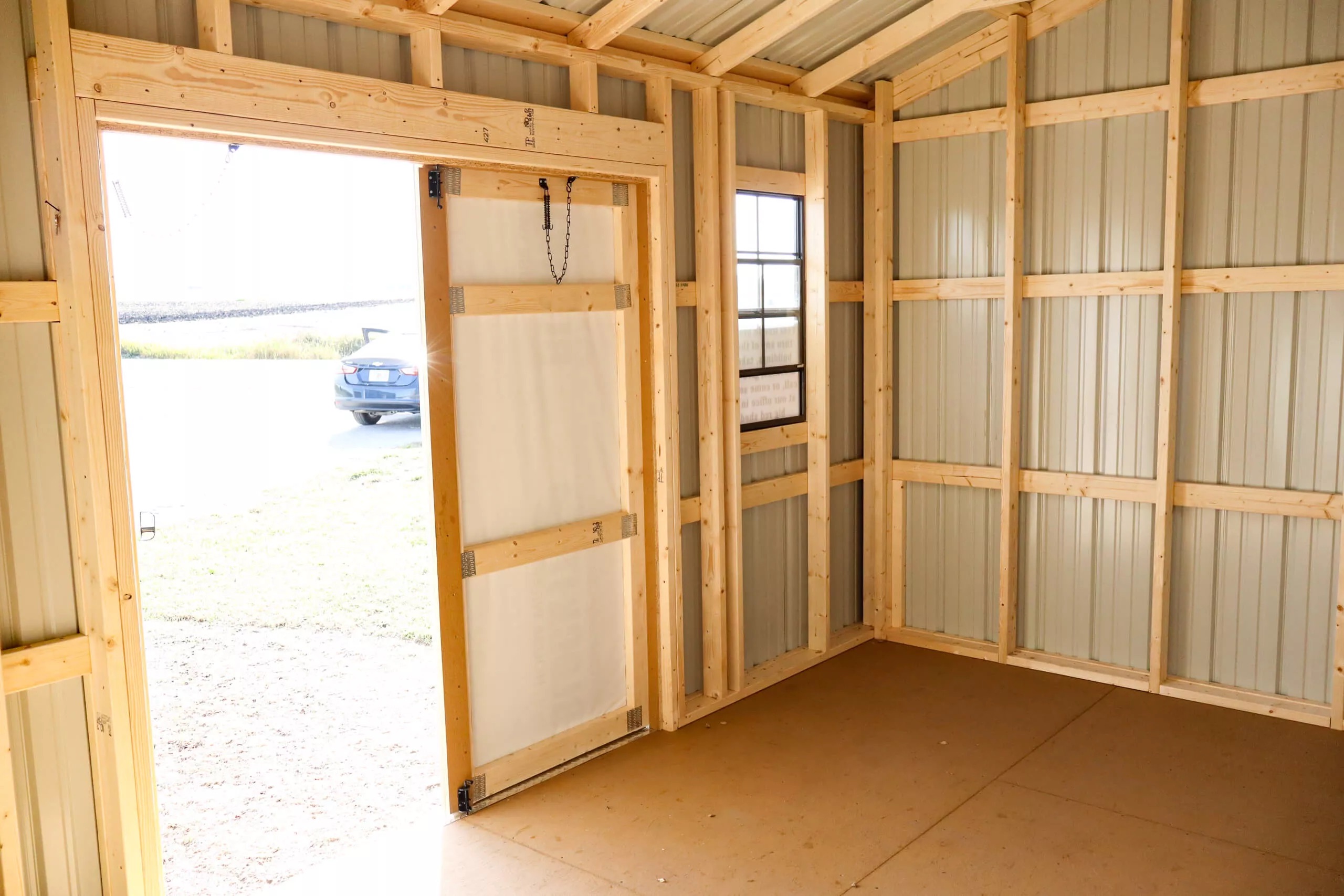The image shows the inside of a small unfinished shed with exposed wooden framing, metal walls, an open door, and a window.