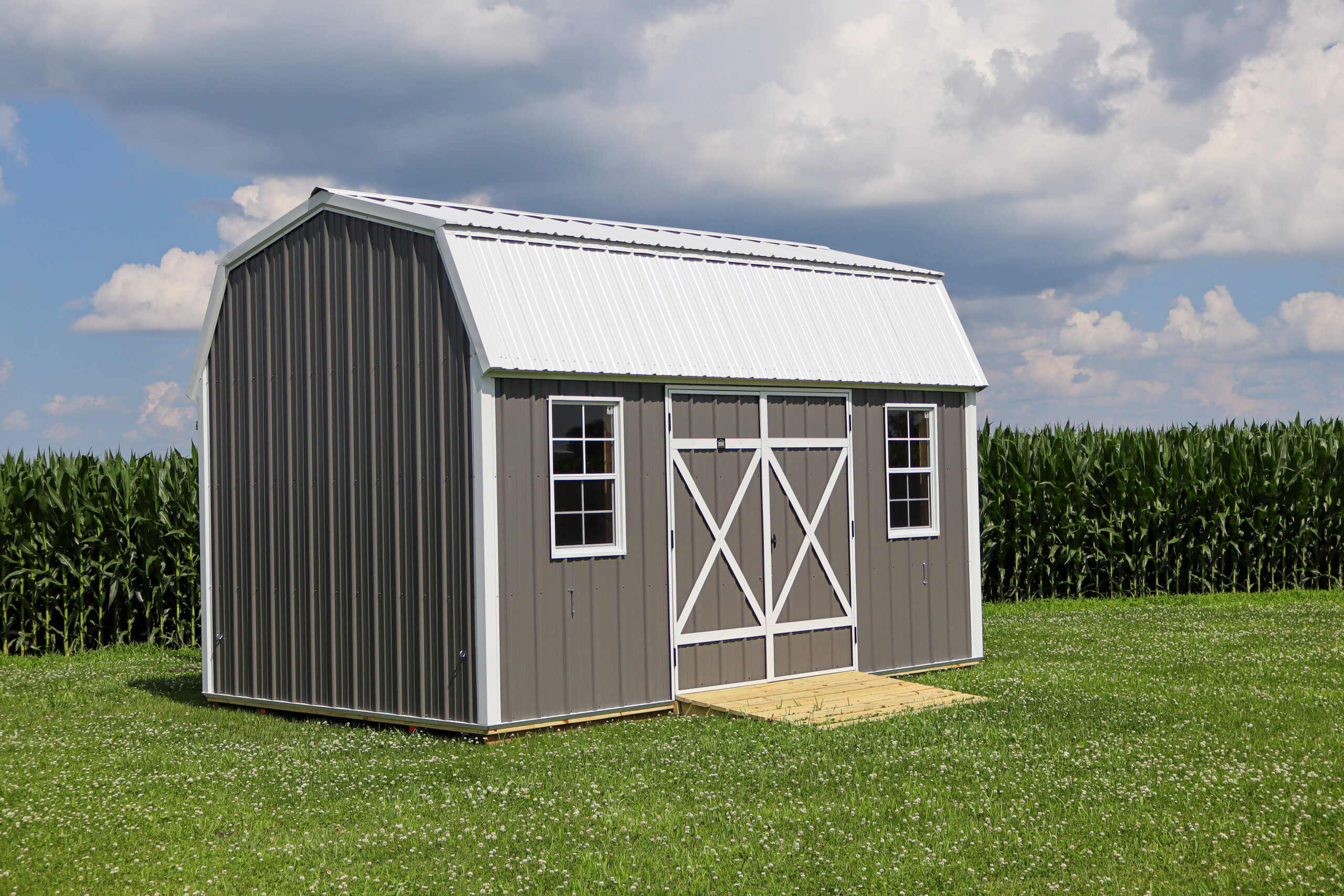 A gray metal shed with white trim and two windows sits on grass in front of a cornfield under a partly cloudy sky.
