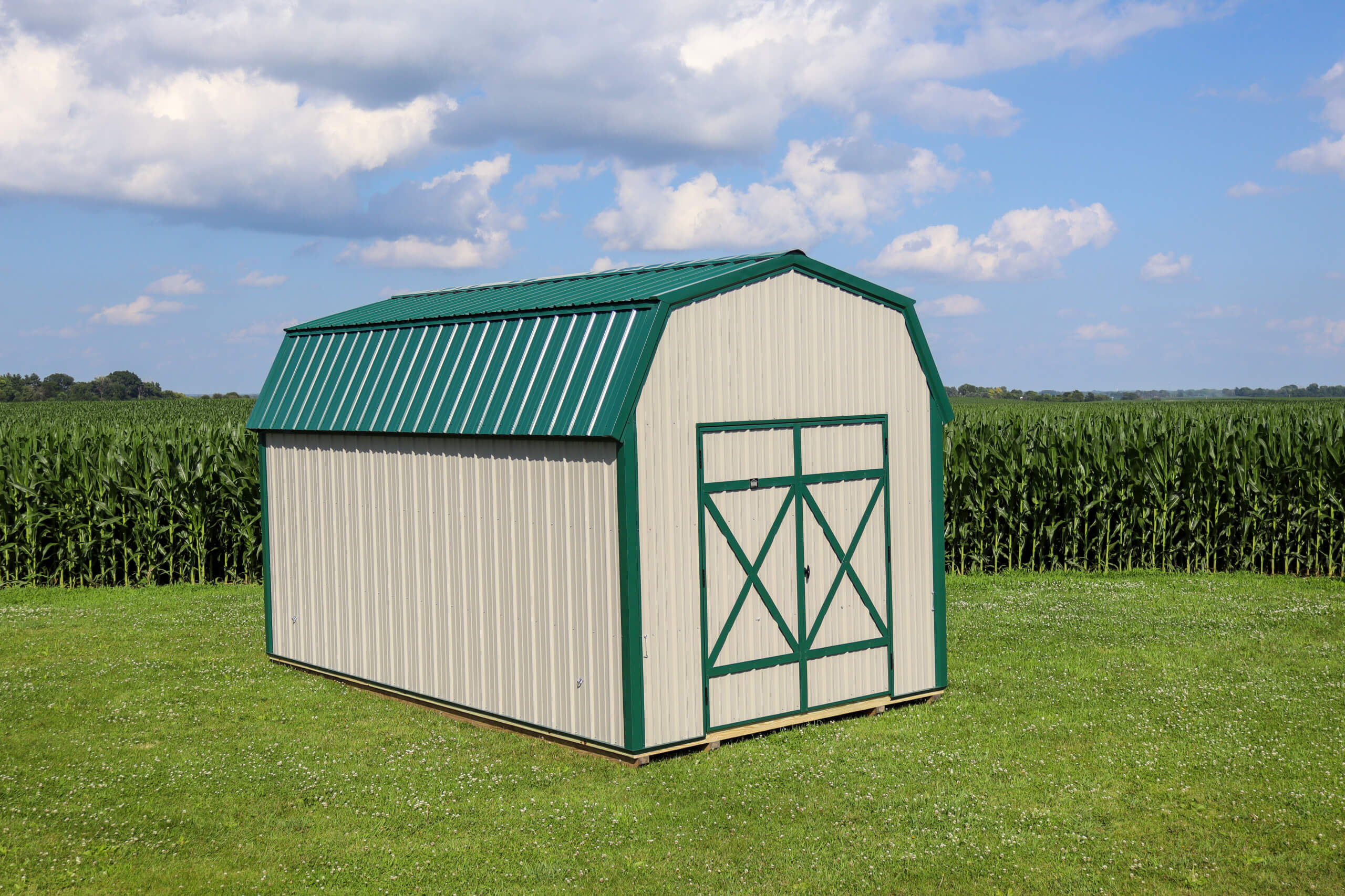 A cream and green metal shed stands on a grassy lawn with a cornfield and blue sky with clouds in the background.