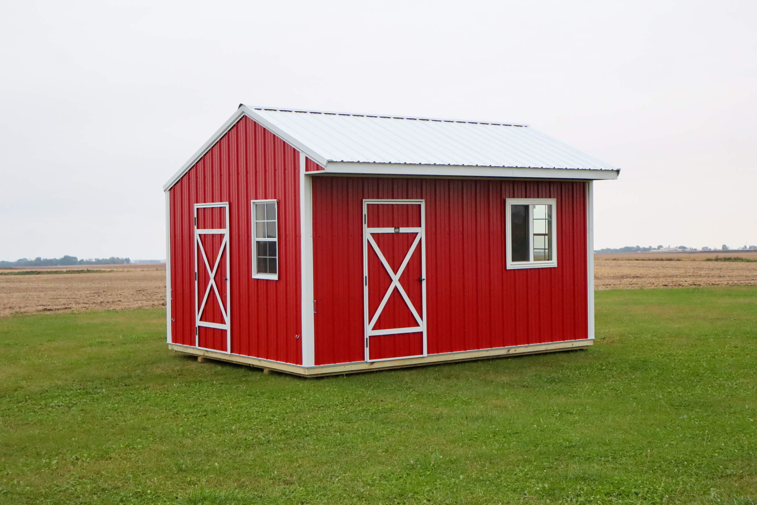 Small red shed with white trim, metal roof, two doors, and a window, sitting on a grassy lawn with fields in the background.