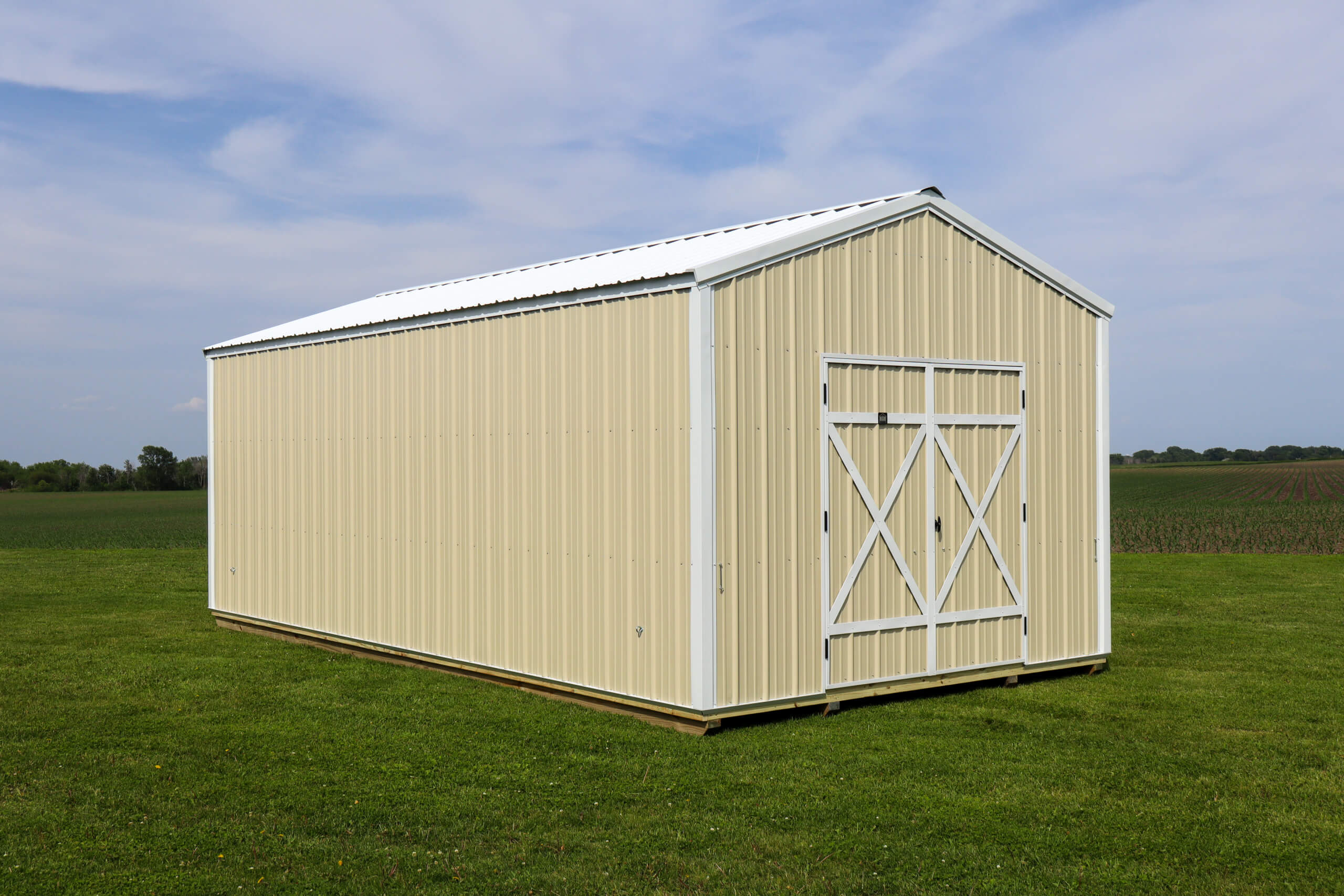 A beige metal storage shed with a white roof and double doors stands on a grassy field under a partly cloudy sky.