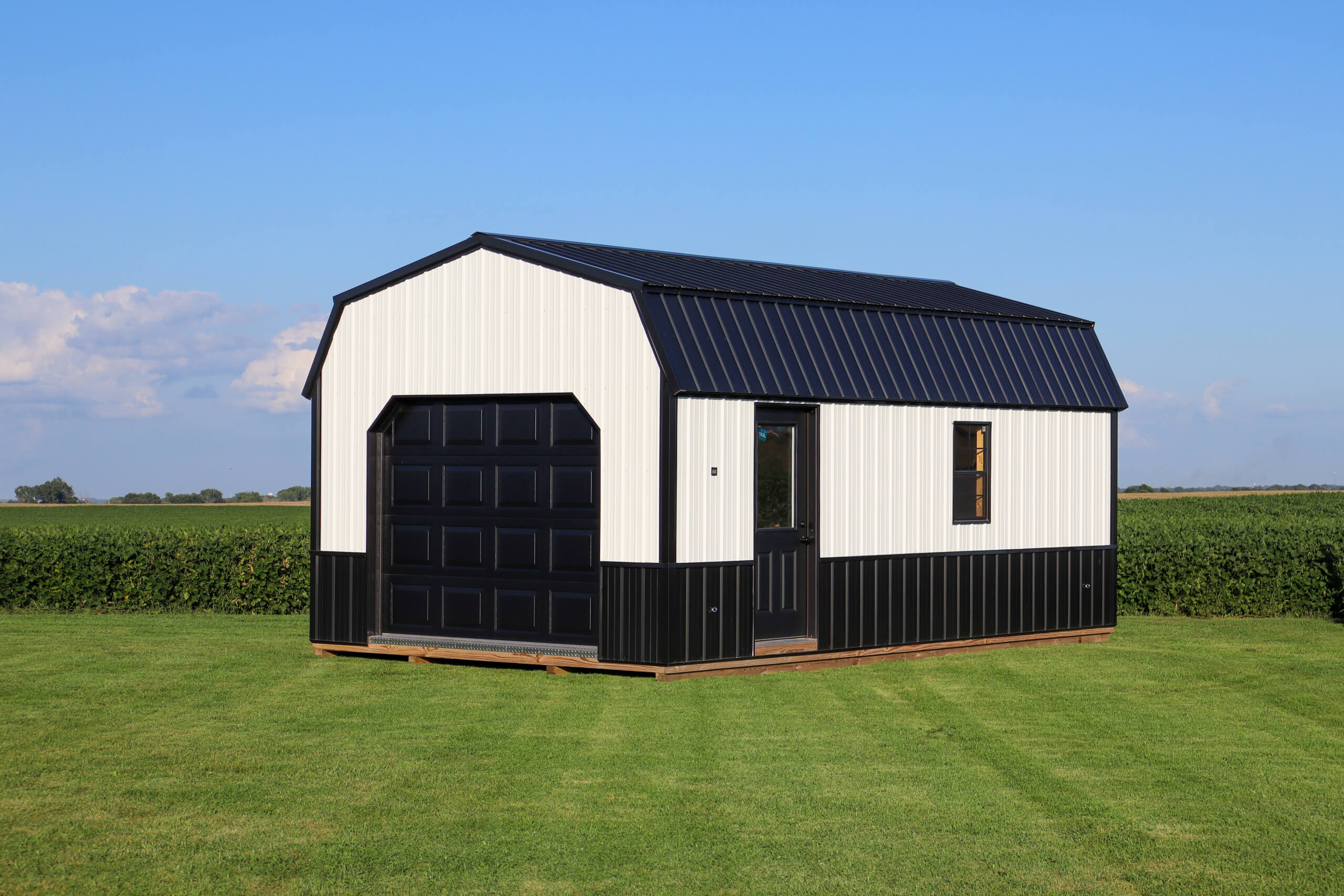 A black and white shed with a garage door and a small window sits on a grassy lawn with farmland in the background under a blue sky.