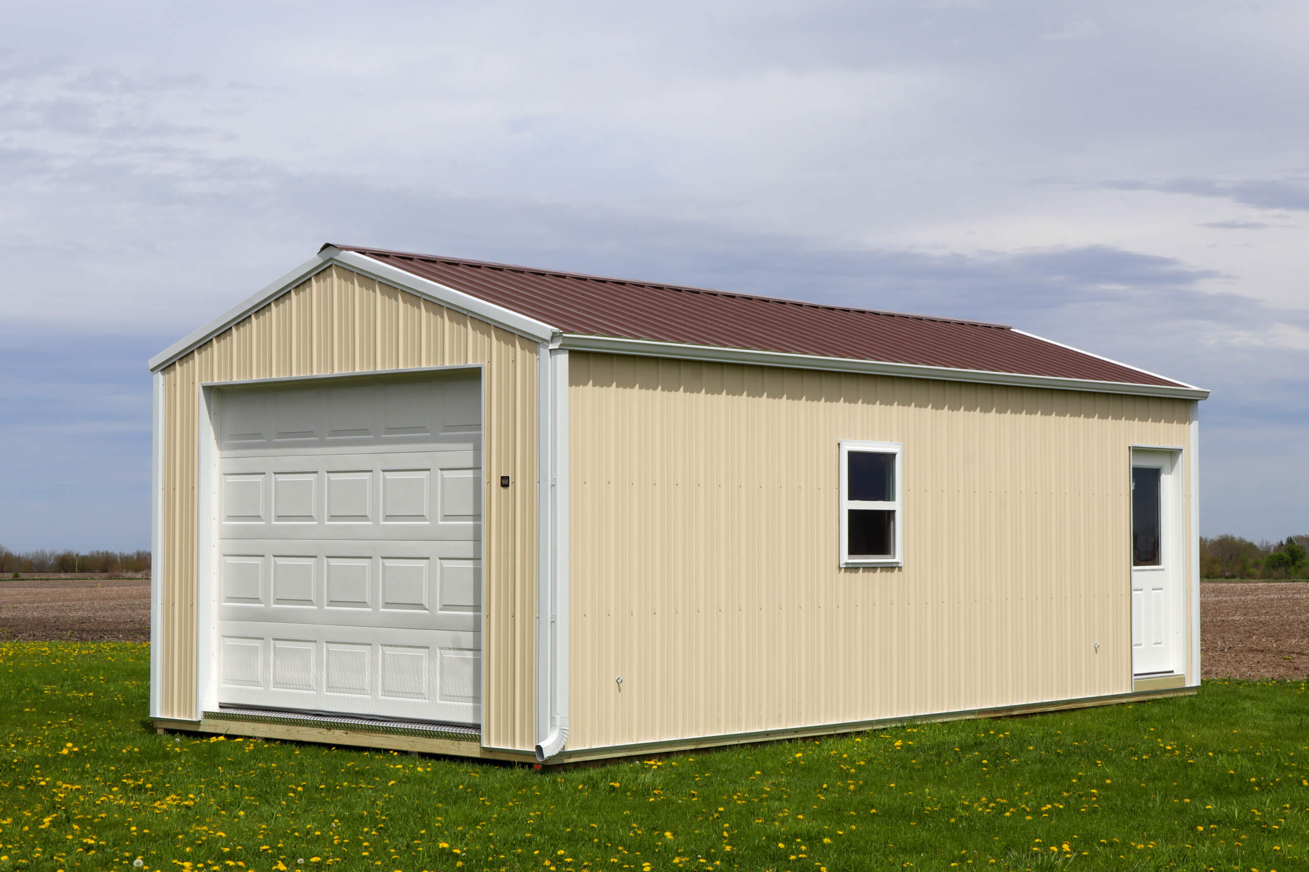 Beige metal shed with a brown roof, white garage door, window, and side door, situated on grass with a cloudy sky in the background.