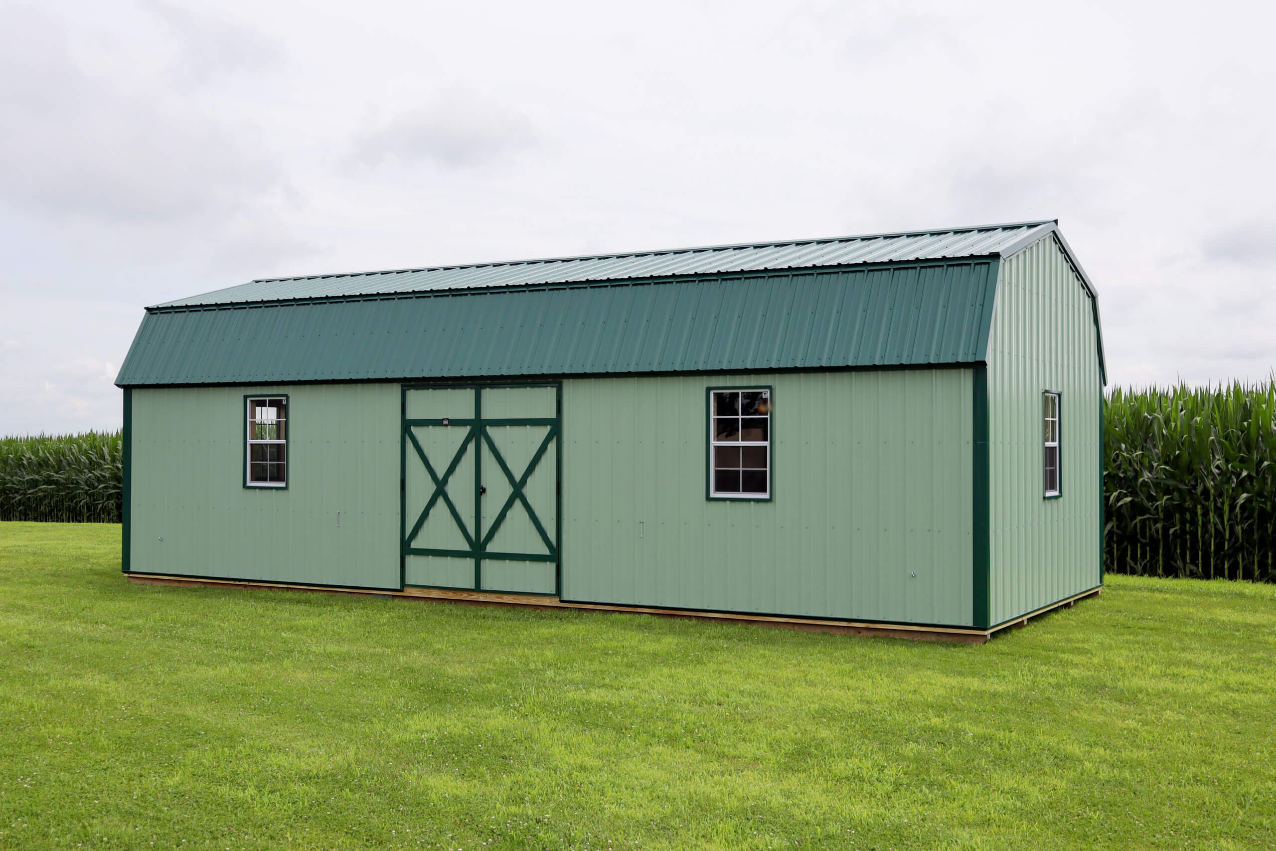 A green metal barn with white trim sits on a grassy field in front of a cornfield under a cloudy sky.